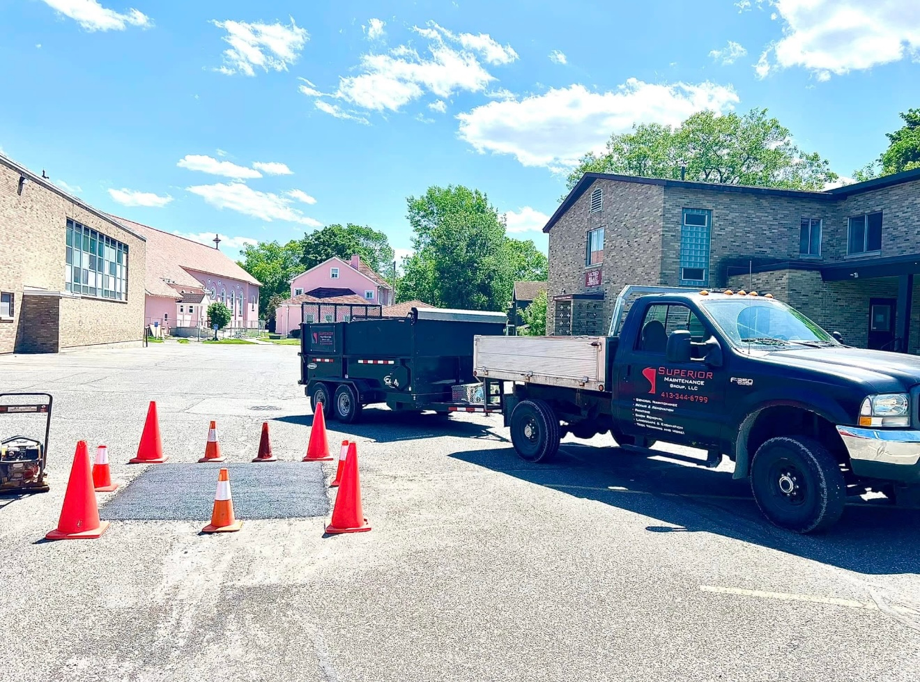 A dark dump truck with an attached trailer sits on a gravel lot near buildings, marked off by several orange safety cones.