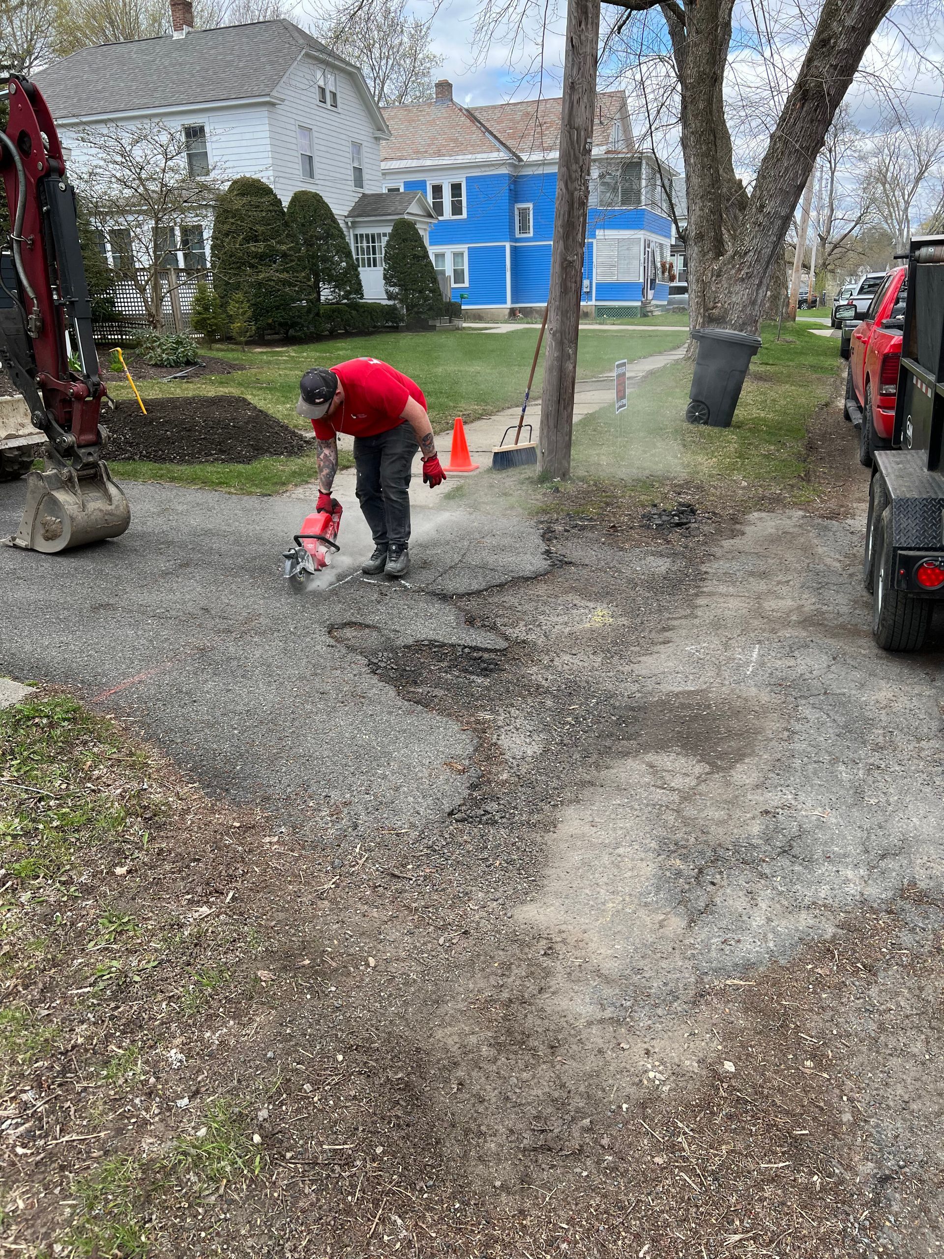 A worker in a red shirt uses a power saw on a gravel driveway in a residential yard near an excavator.