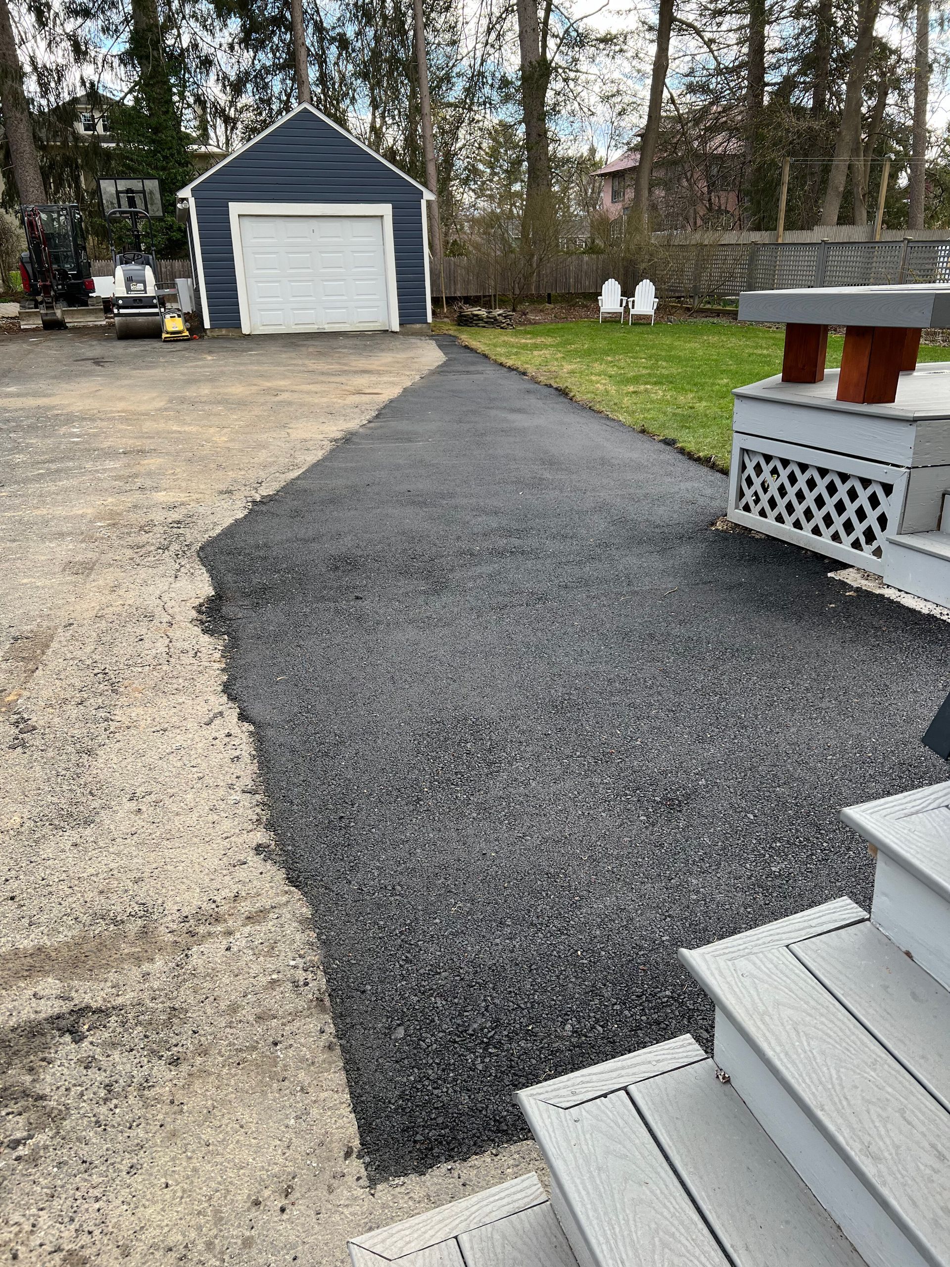 A newly paved asphalt walkway leading from wooden deck steps toward a blue garage in a backyard setting.