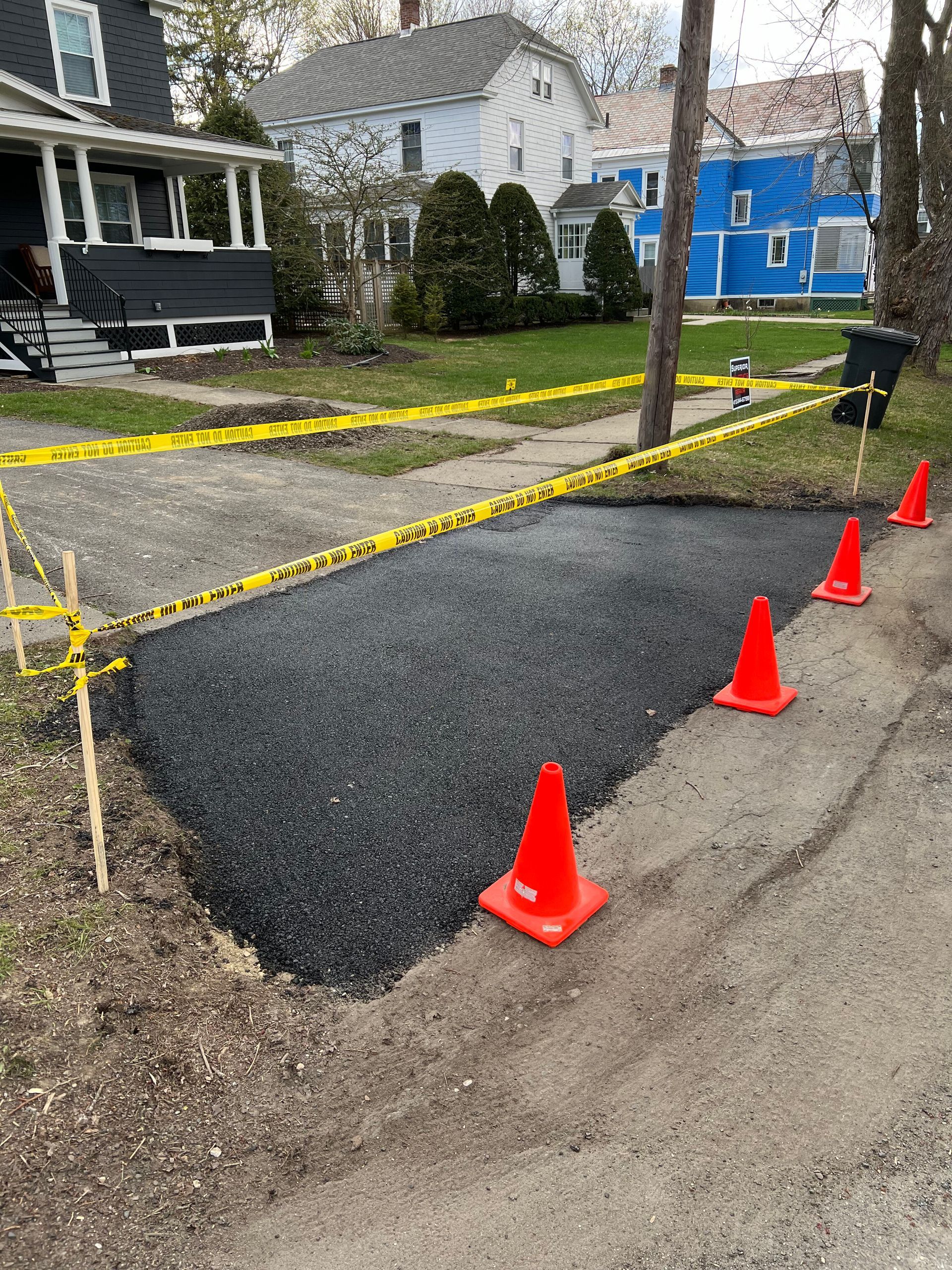 Freshly laid black gravel fills a driveway section, cordoned off by yellow caution tape and orange traffic cones.