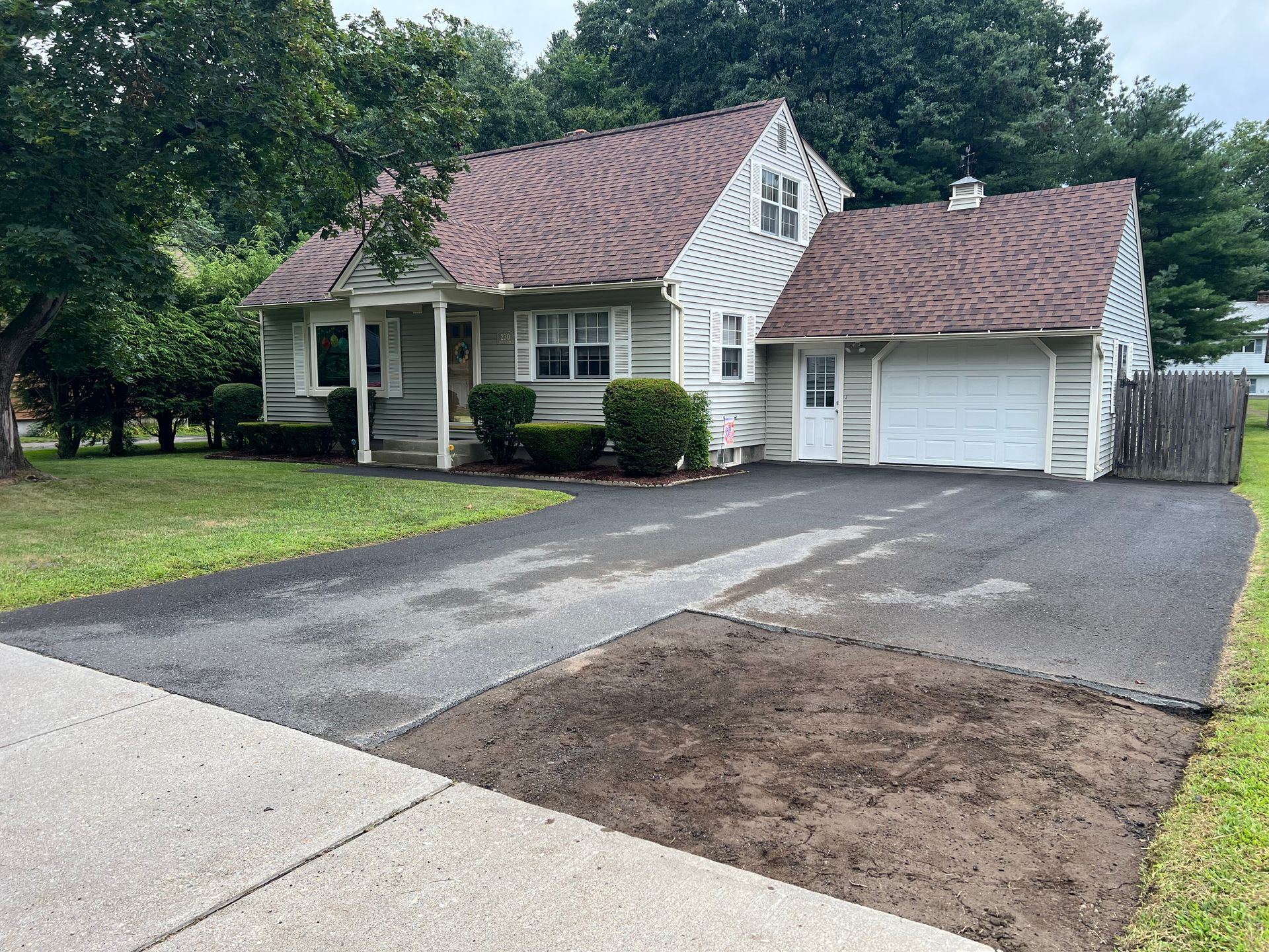 A light-colored house with a dark roof and an attached garage, featuring a driveway with a rectangular patch of dirt.