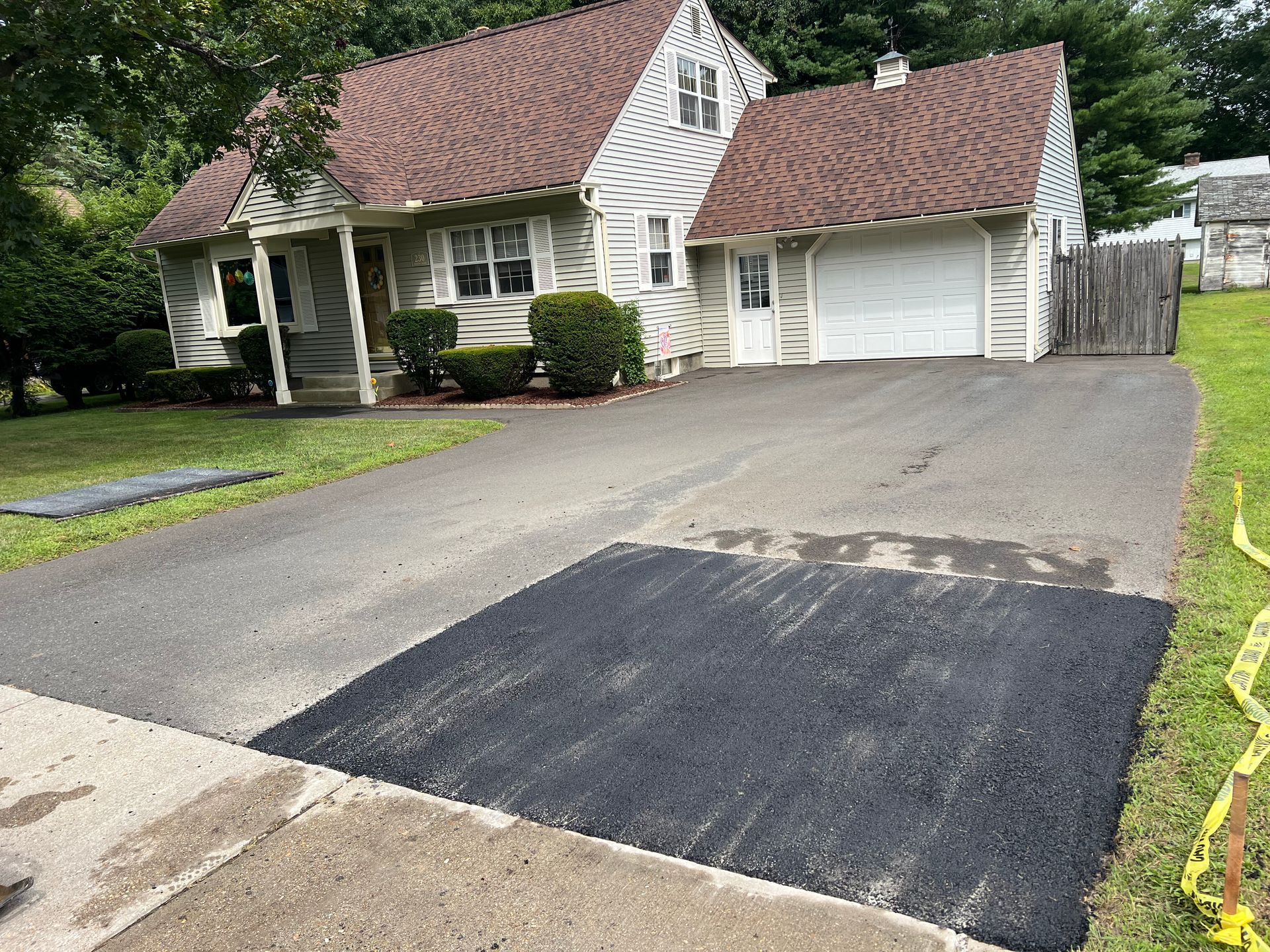 A suburban house with brown roofing and a white driveway featuring a freshly paved, dark asphalt rectangular patch.