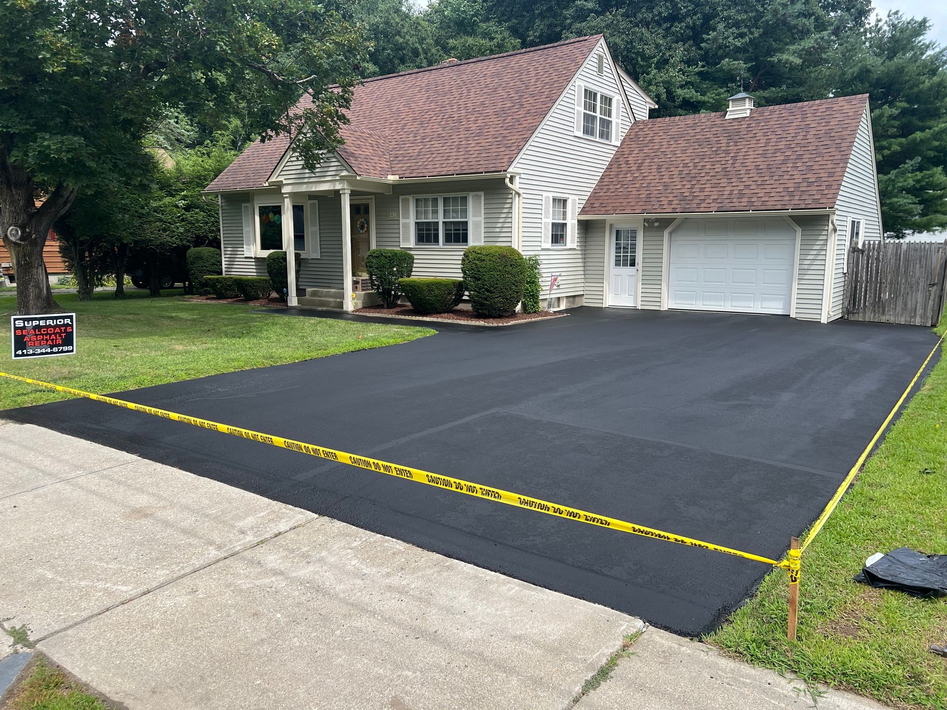 A newly paved black asphalt driveway in front of a house, blocked off with yellow caution tape on a residential lawn.