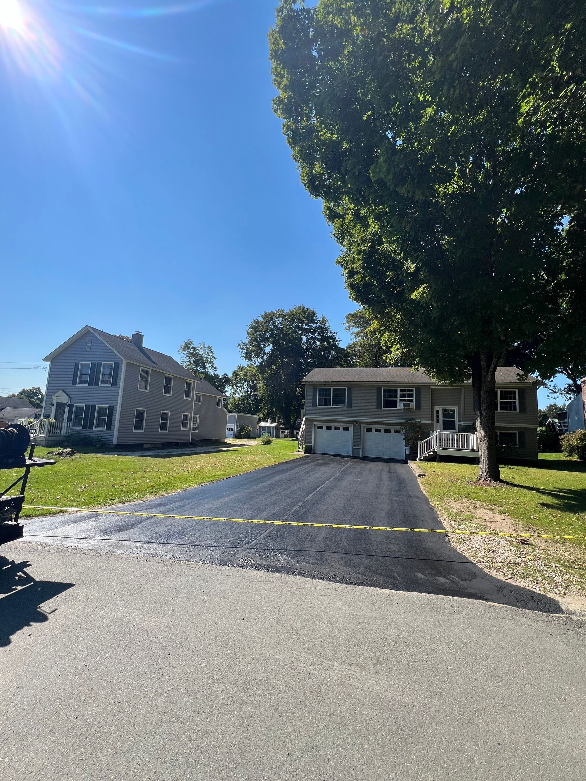 A freshly paved, dark asphalt driveway leads to a suburban two-story house under a bright blue, sunny sky.
