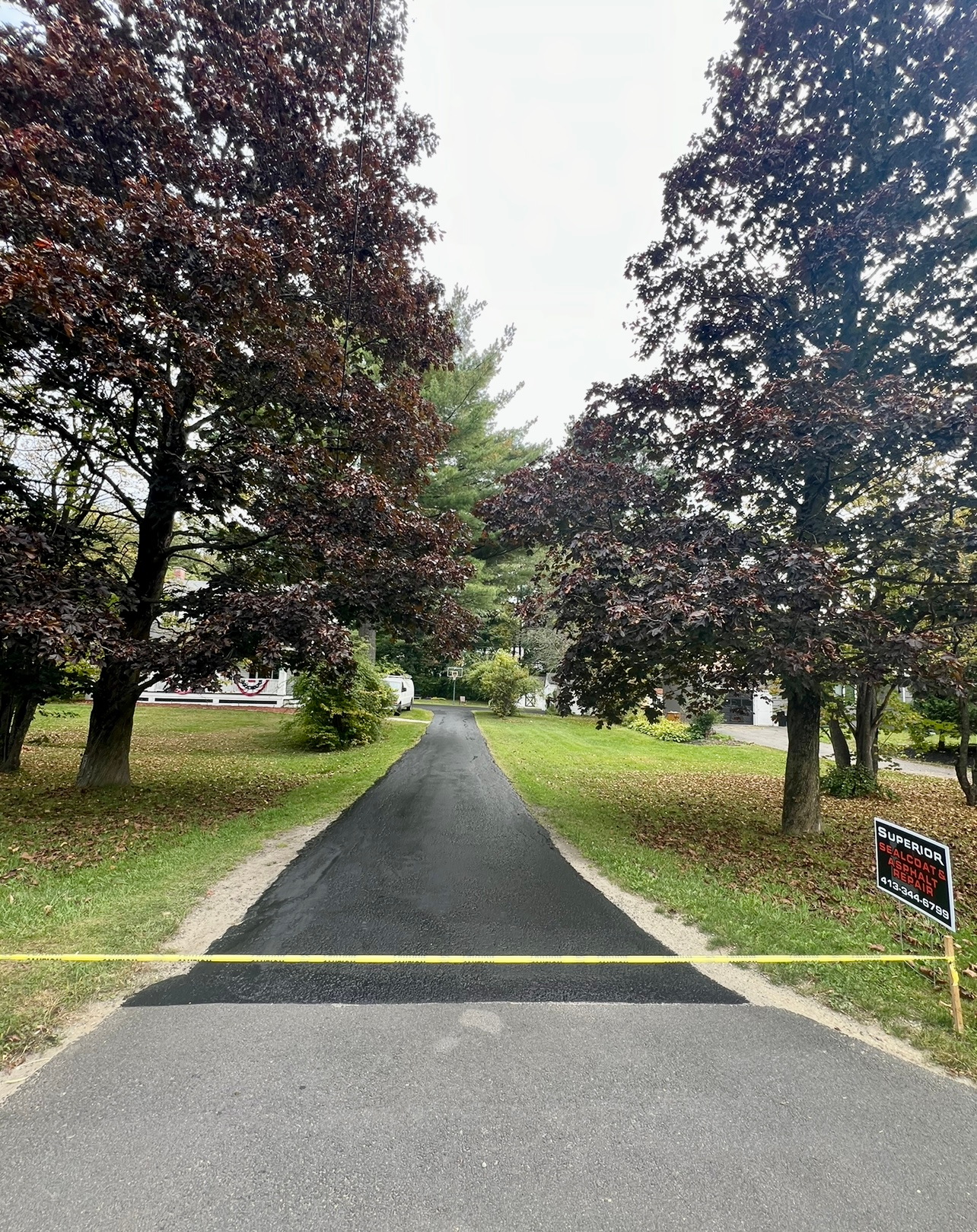 A freshly paved black asphalt driveway stretches between two large trees toward a house, bisected by a yellow caution tape.
