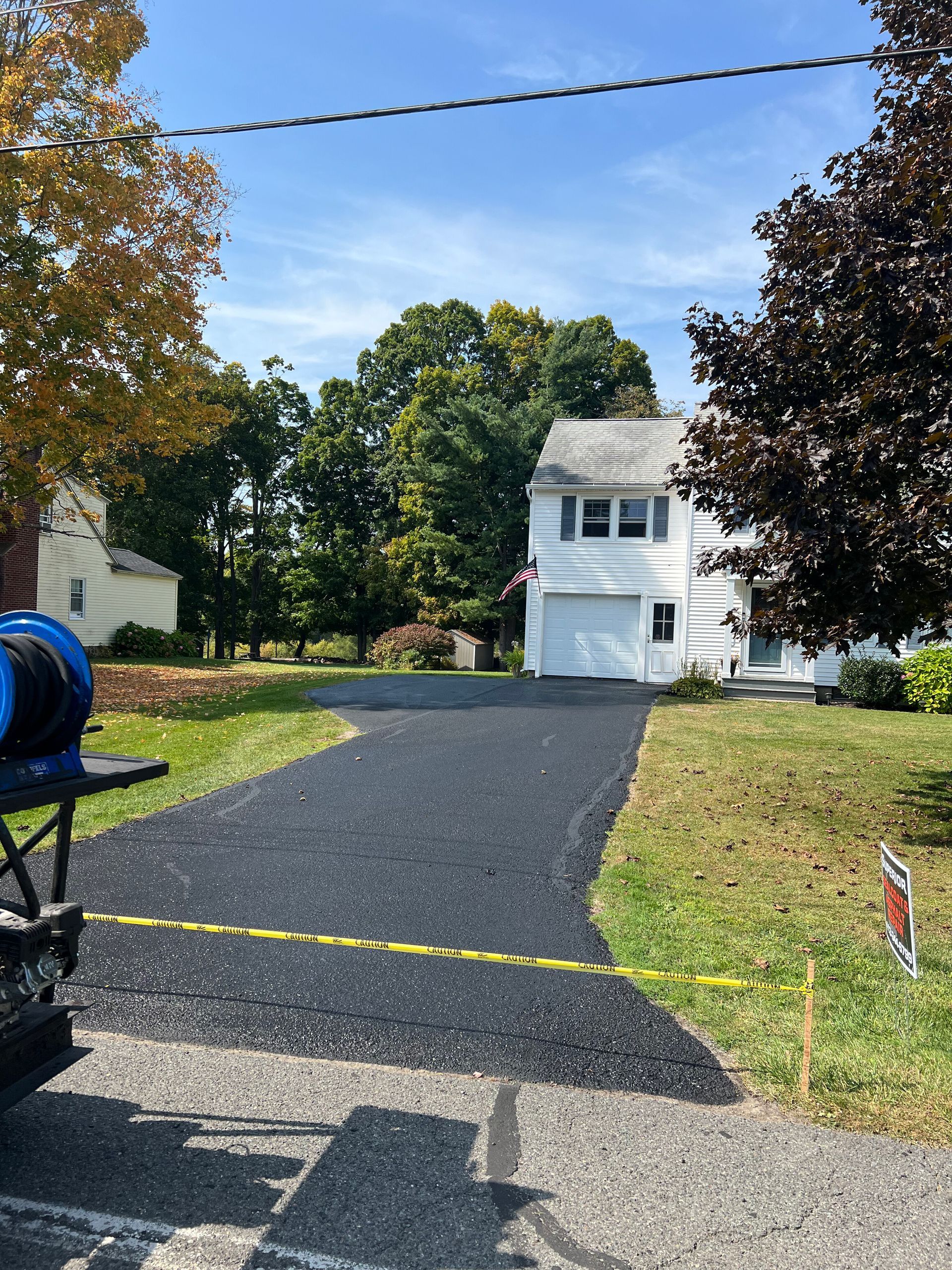 A freshly paved black asphalt driveway leads to a white house with a garage, under a clear blue sky on a sunny day.