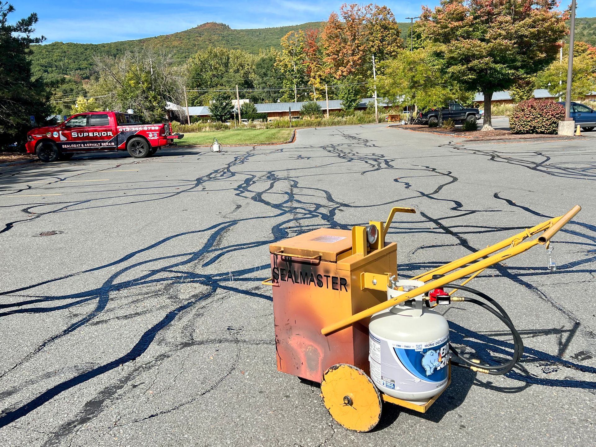 A yellow propane-powered asphalt crack melter sits on a paved parking lot with black sealant lines and a red pickup truck.