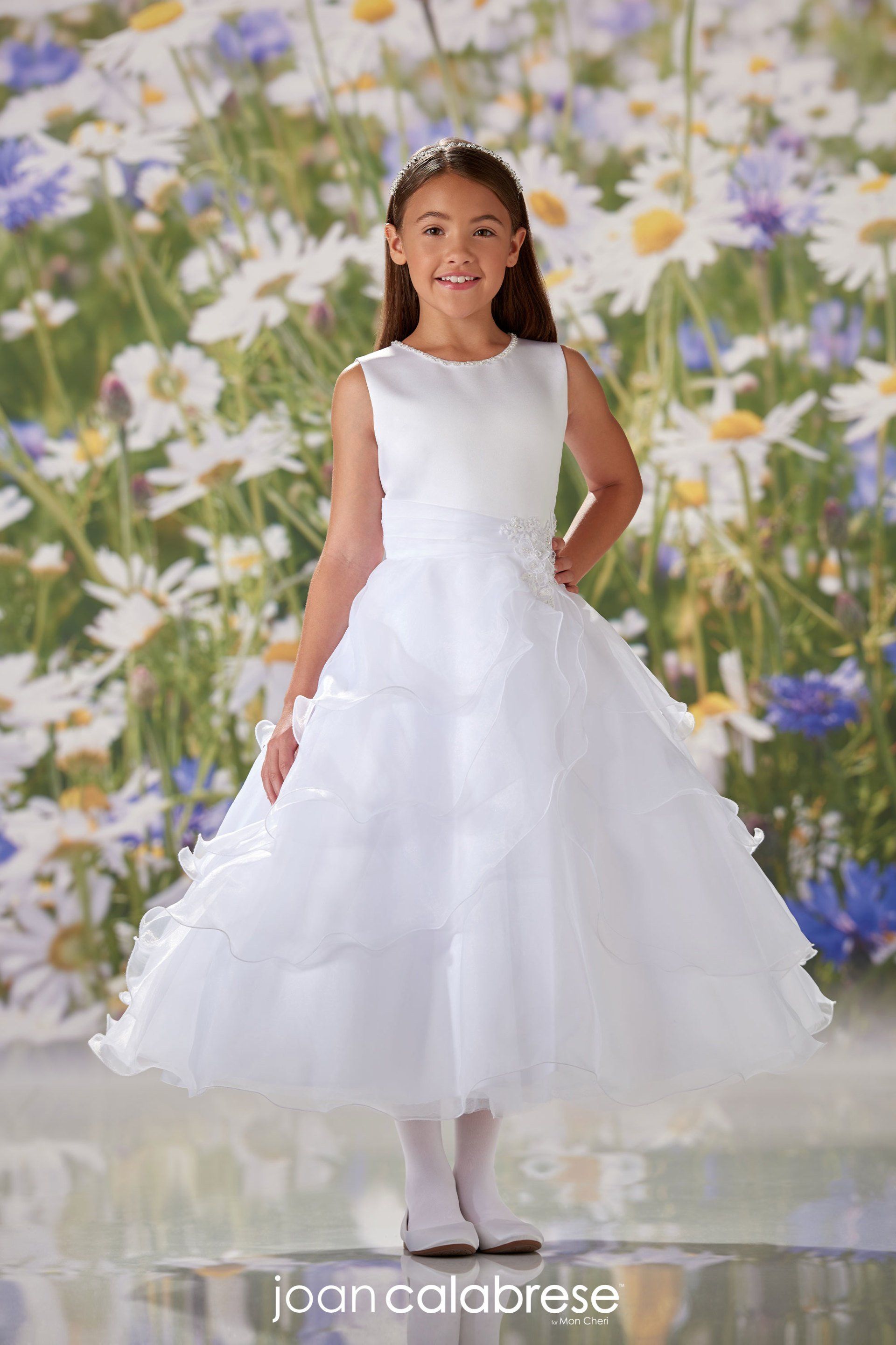 Young girl in a white dress standing before a floral backdrop, smiling with hands behind her back