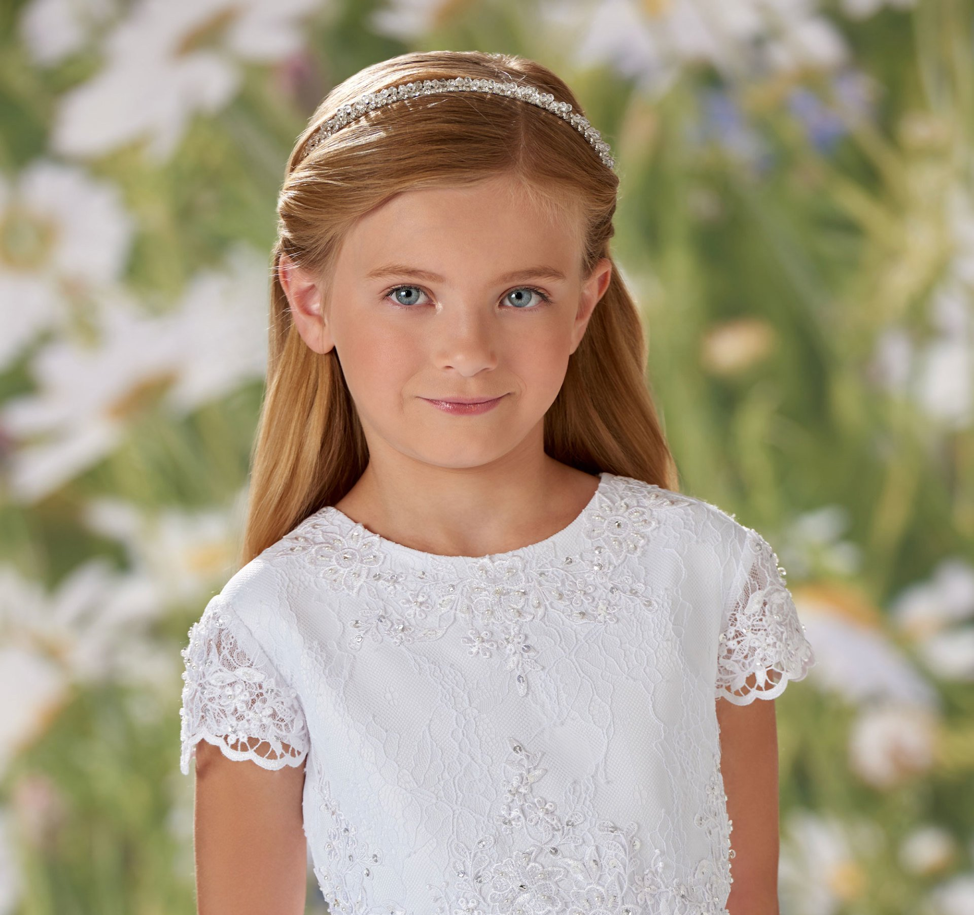 Young girl in a white lace dress standing outdoors with a soft floral background