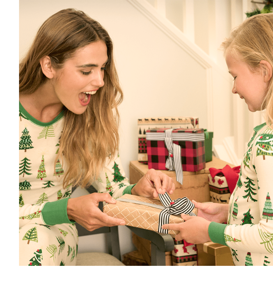 Two people in matching holiday pajamas wrapping a gift by a decorated Christmas tree