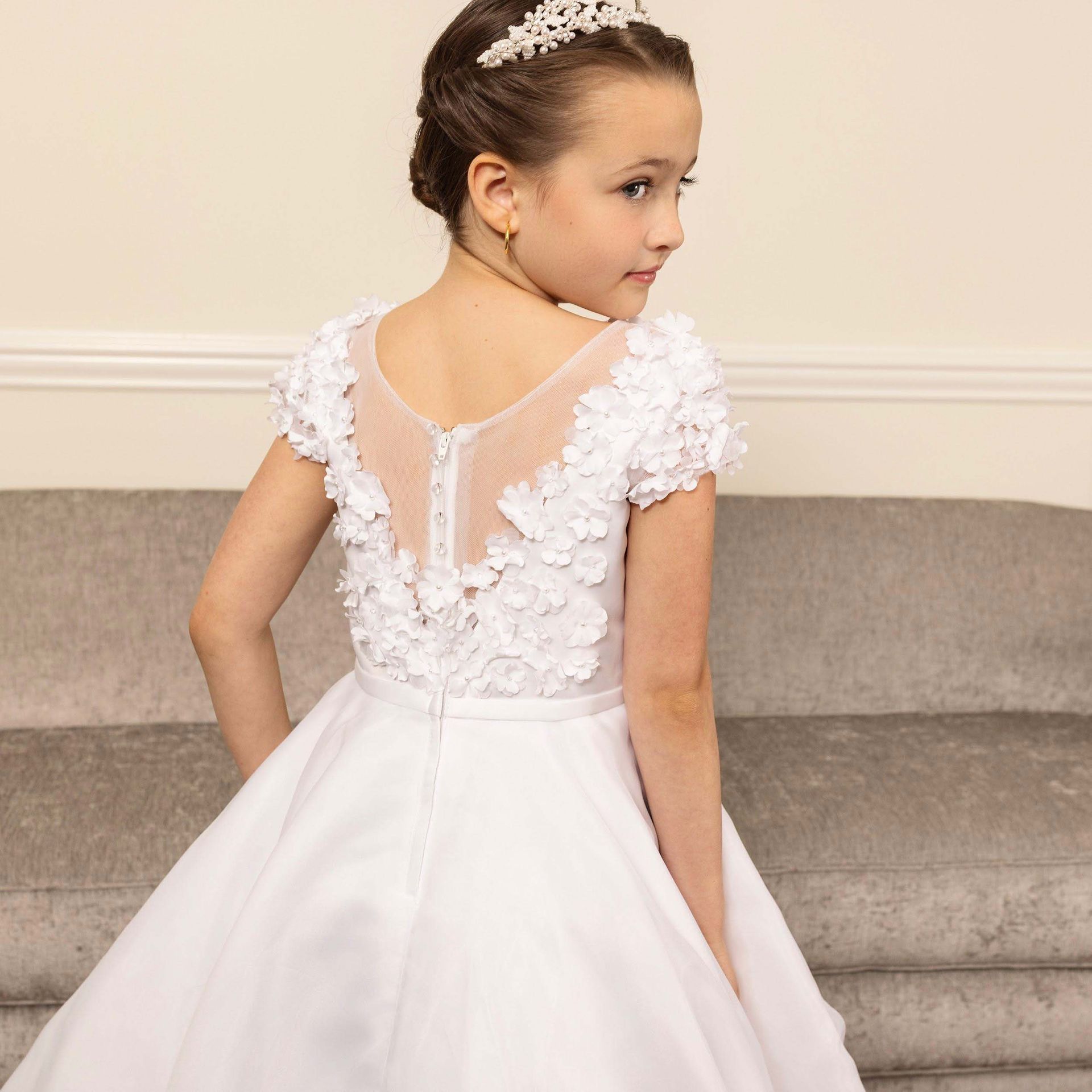 Young girl in a white formal dress with lace back details, seated on a gray couch and looking over her shoulder