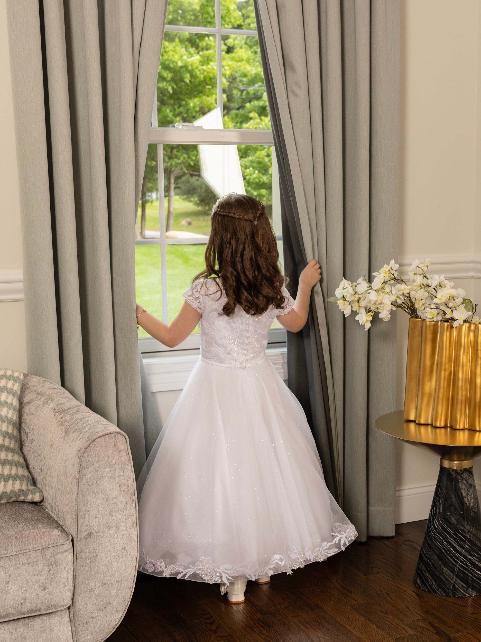 Girl in a white dress stands at a window, looking out through gray curtains in a bright room.
