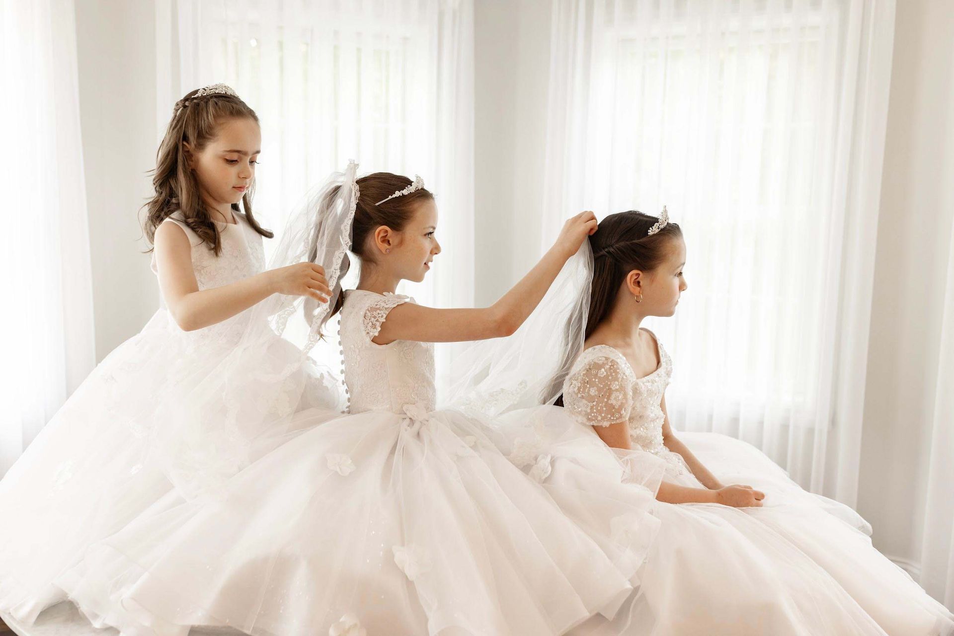 Three children in white dresses adjusting each other's hair by a bright window