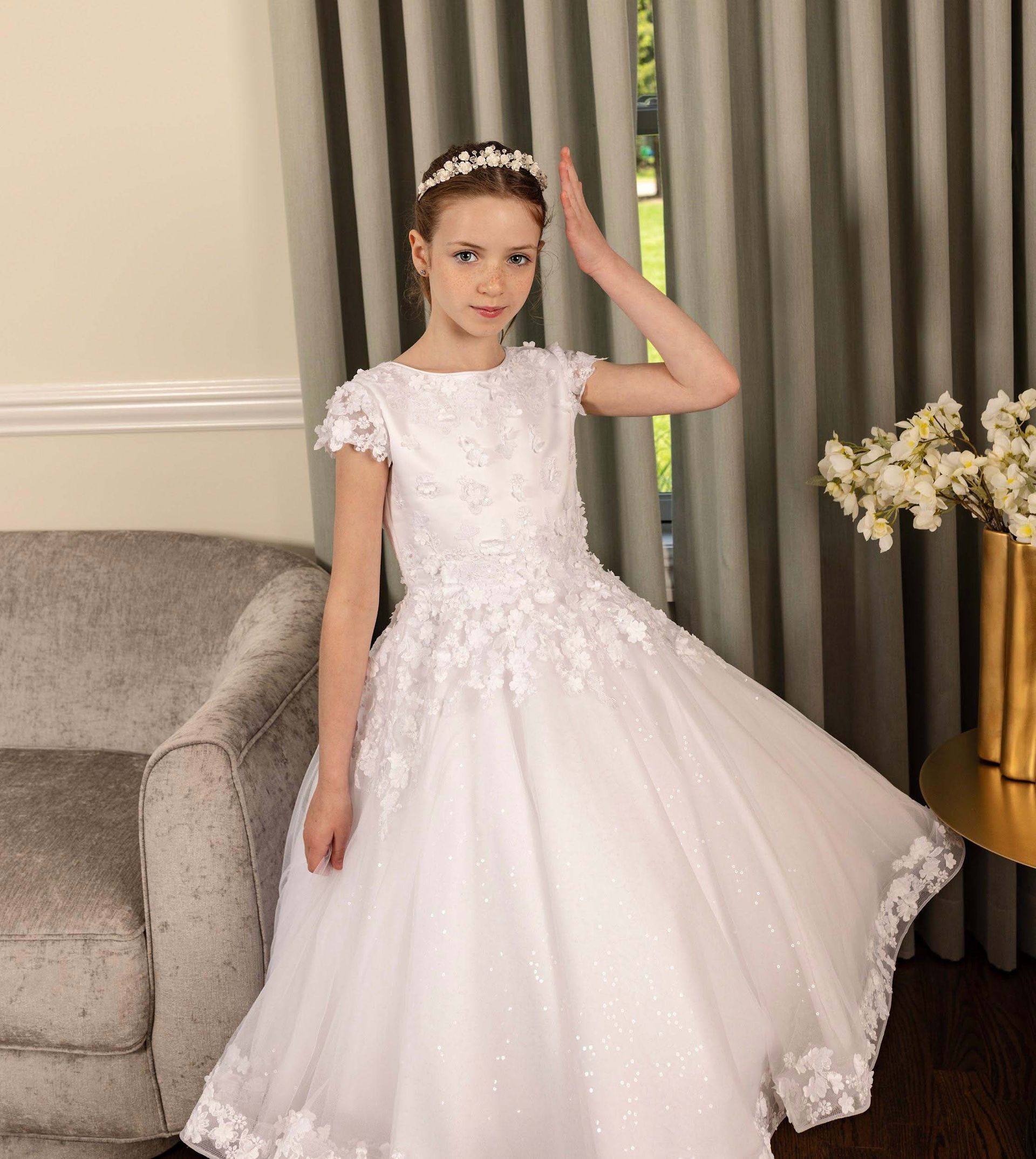 Young girl in a white formal dress standing beside a sofa and curtains indoors