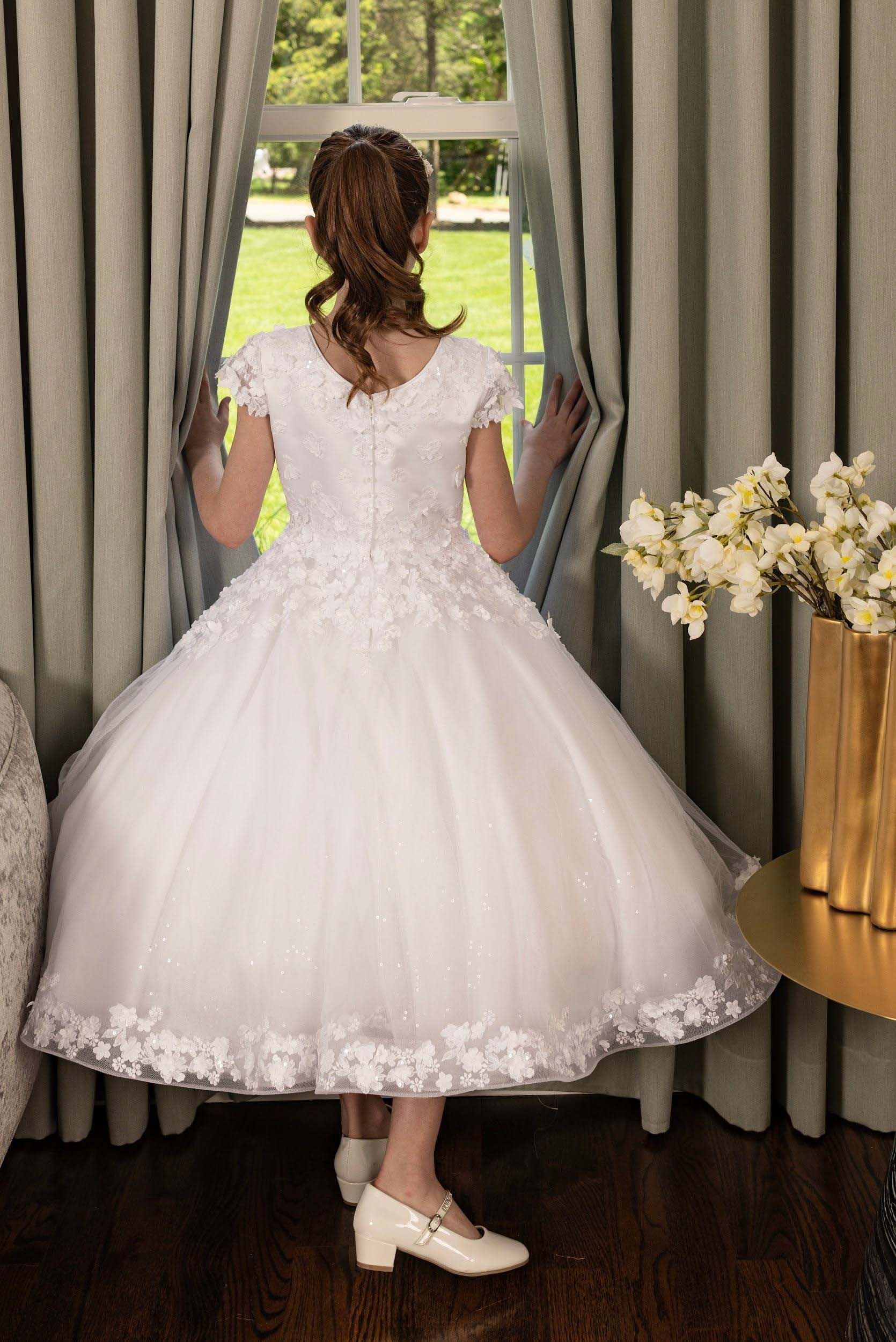 Girl in a white dress standing by a window, facing away, with curtains and flowers beside her.