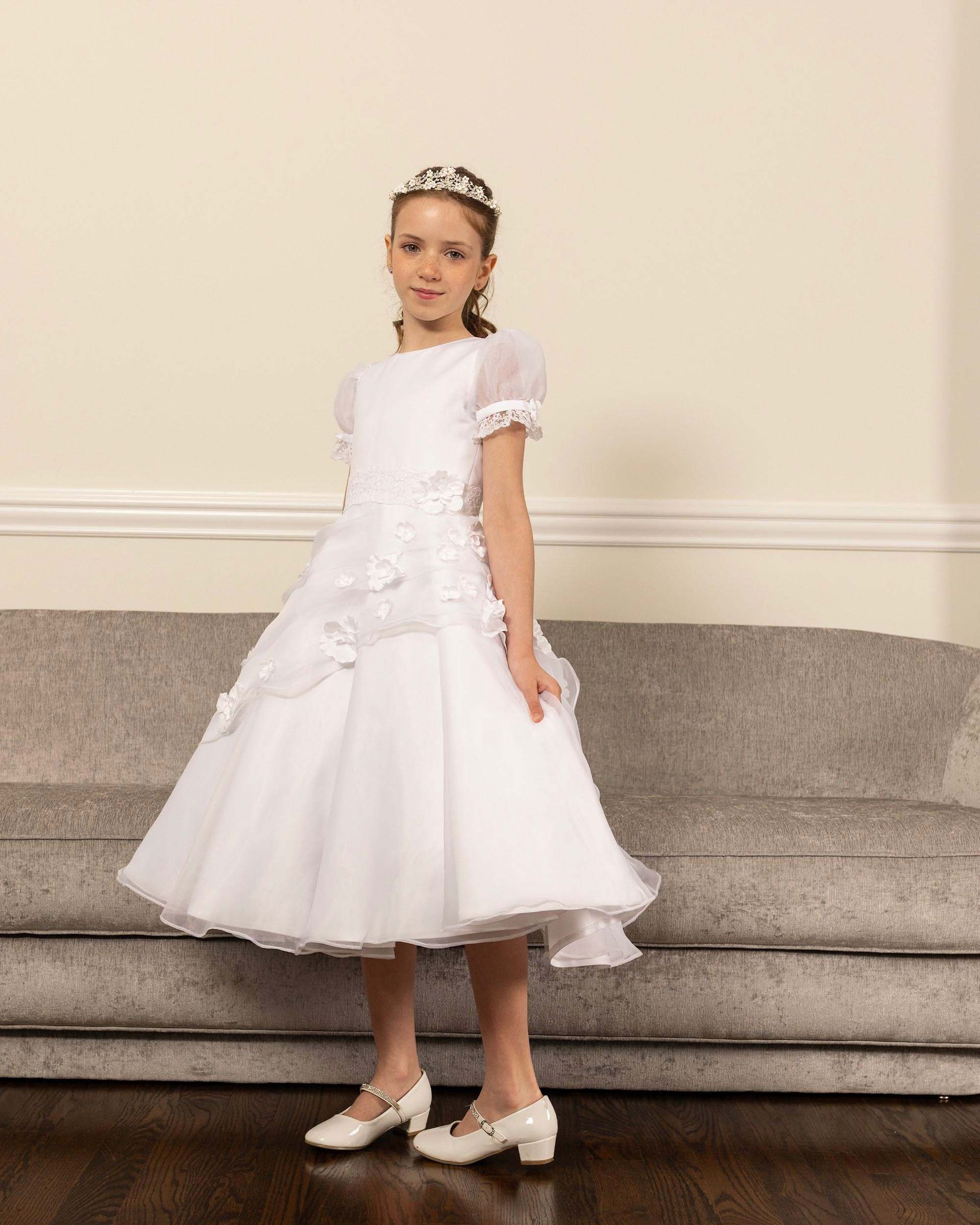 Young child in a white dress and headband standing in a living room beside a beige sofa