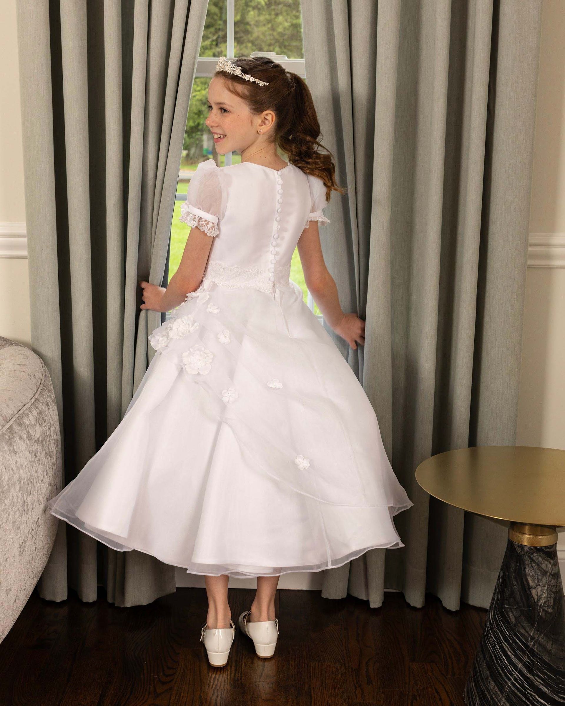Girl in a white dress standing by curtains, looking over her shoulder in a formal room.