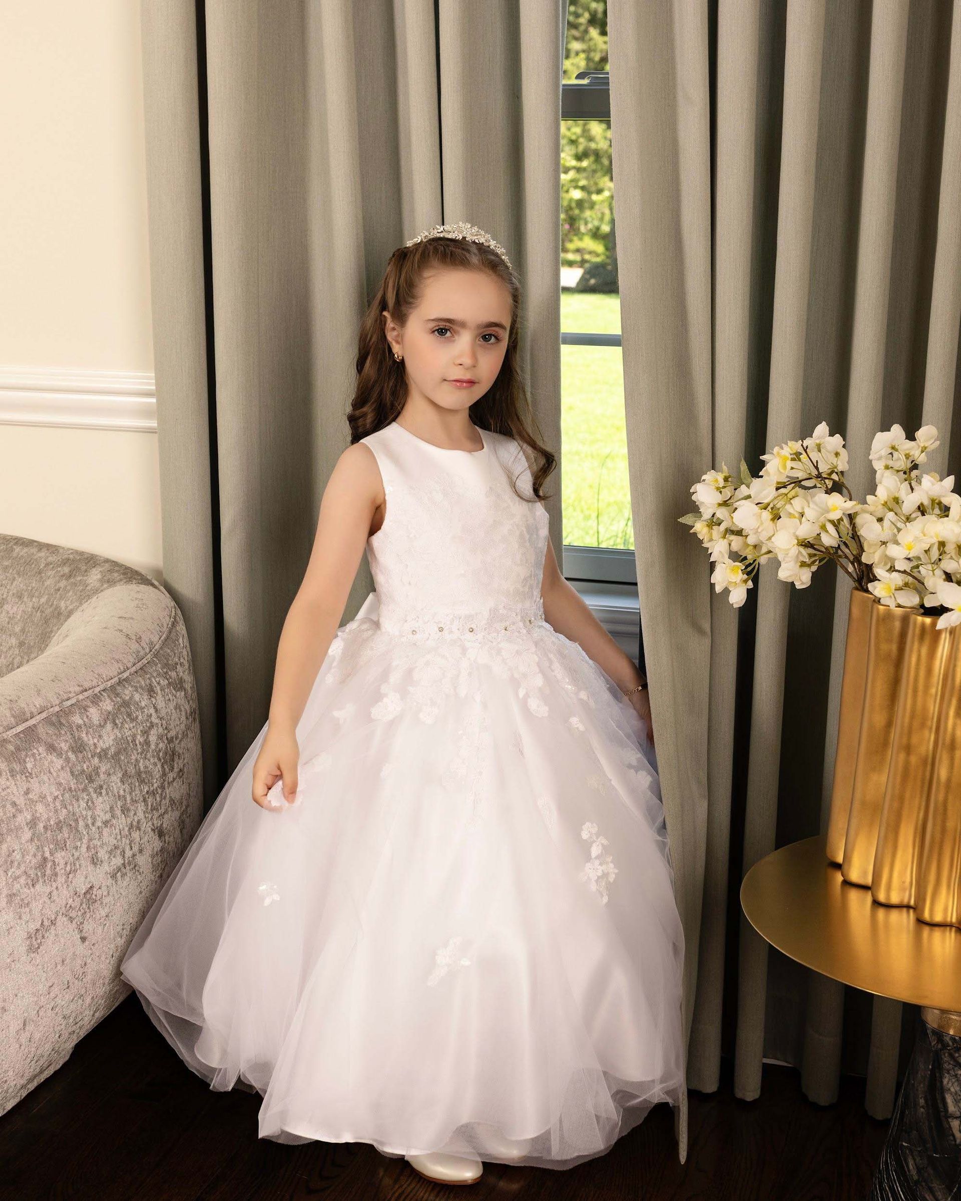 Young child in a white tulle dress standing indoors beside curtains and a vase of flowers