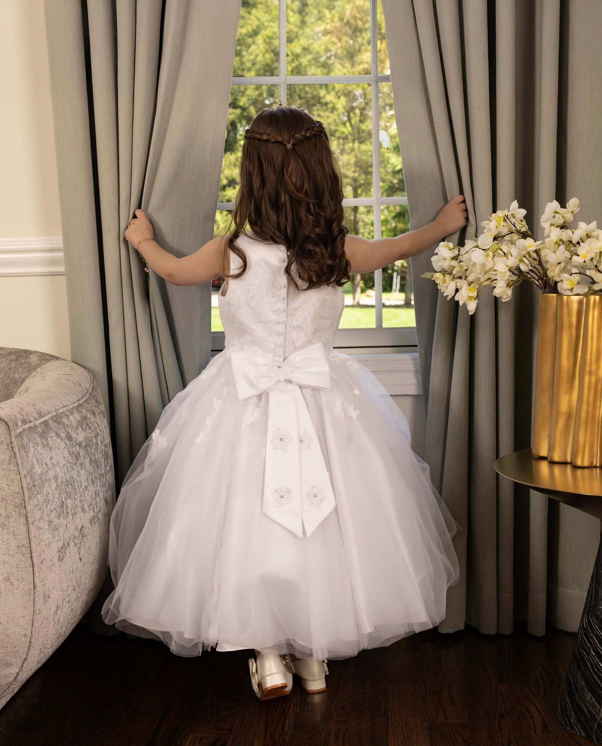 Young child in a white dress standing by a window, holding open gray curtains in a bright room.