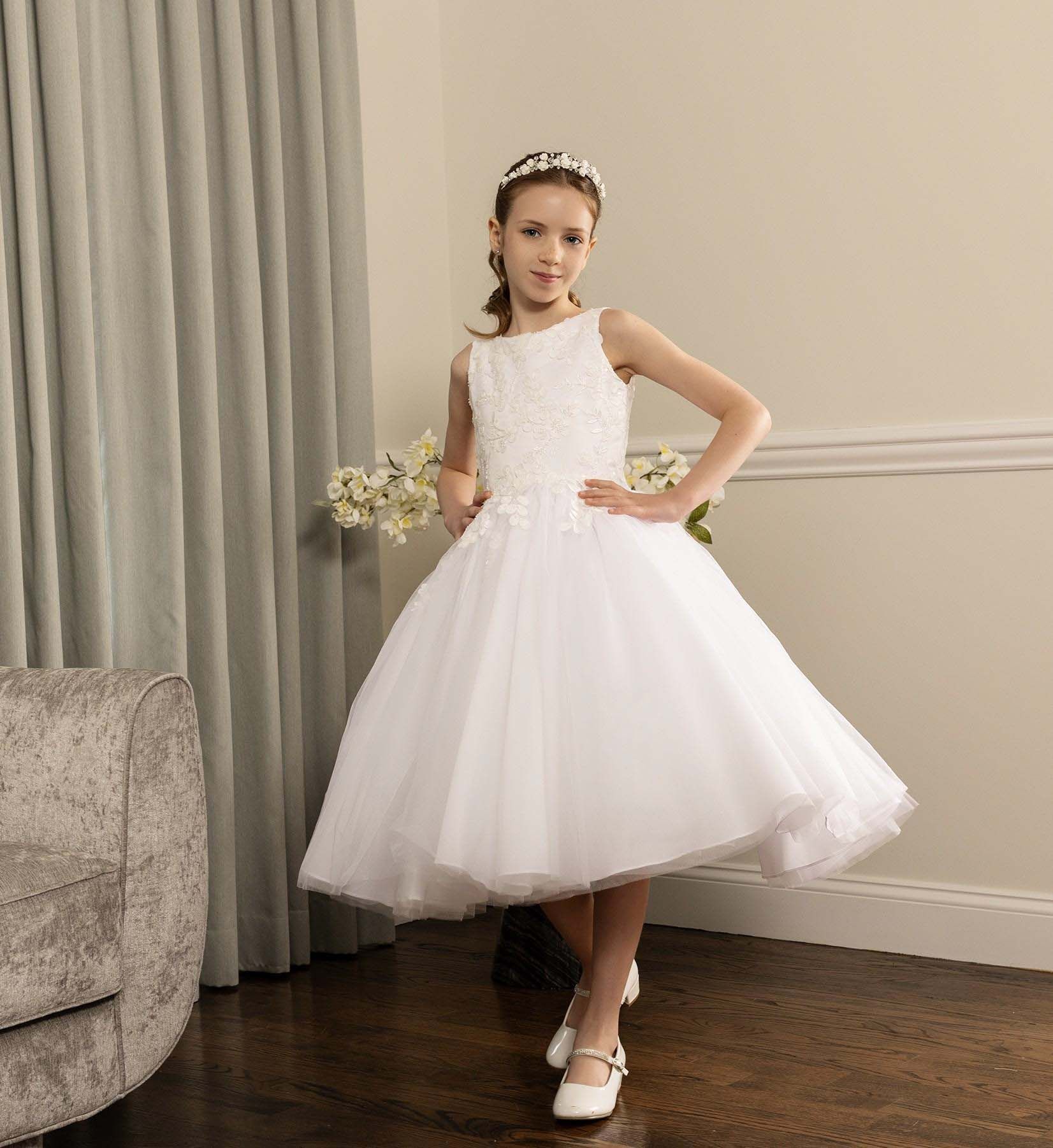 Girl in a white dress posing indoors on a hardwood floor, holding flowers.