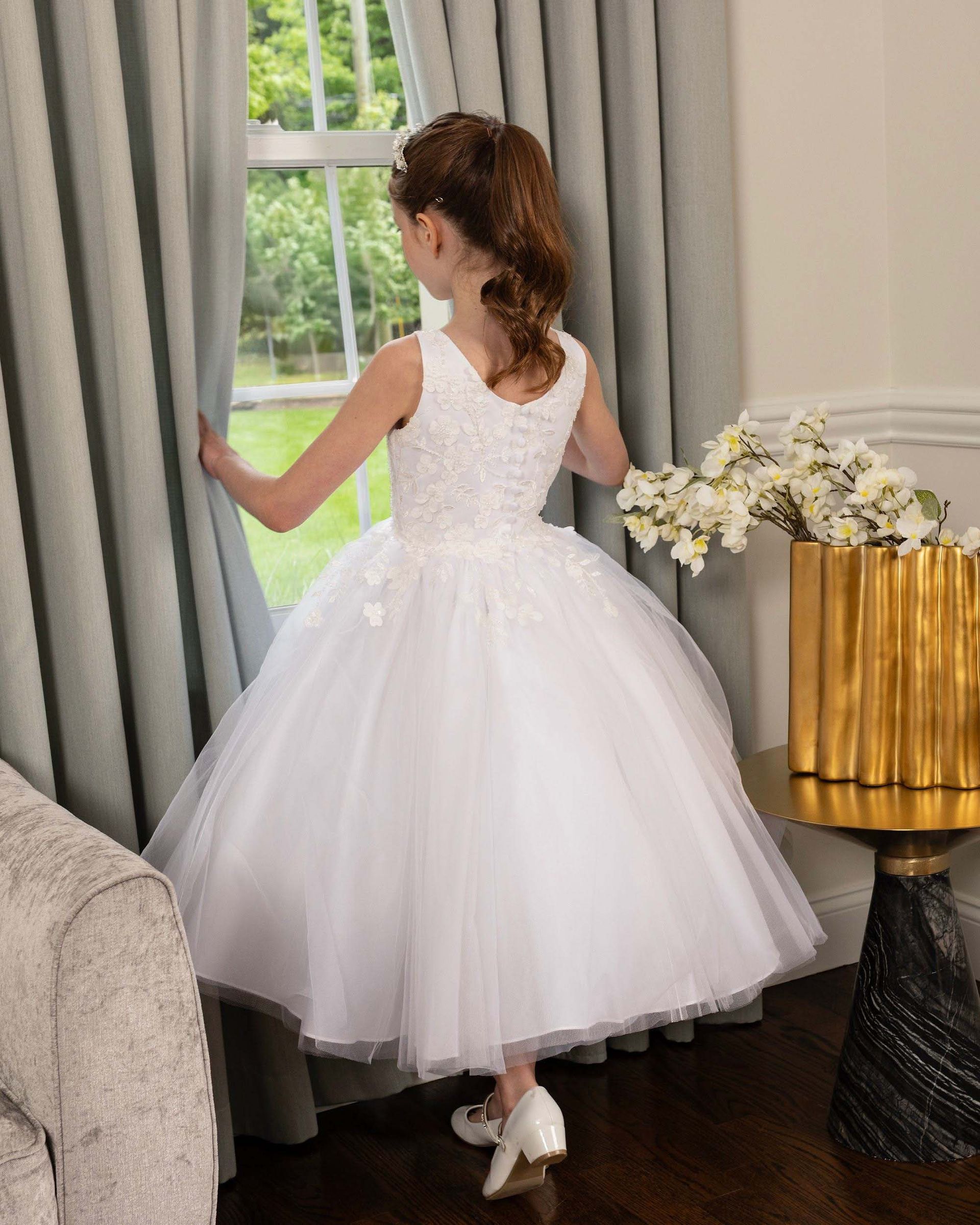 Girl in a white dress standing by a window, holding curtains in a bright living room.