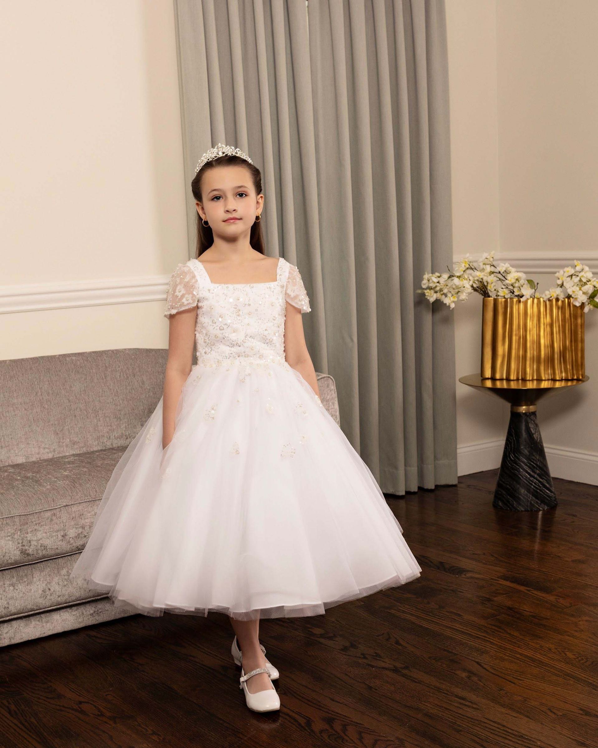 Child in a white dress and tiara stands indoors beside a staircase and curtain.