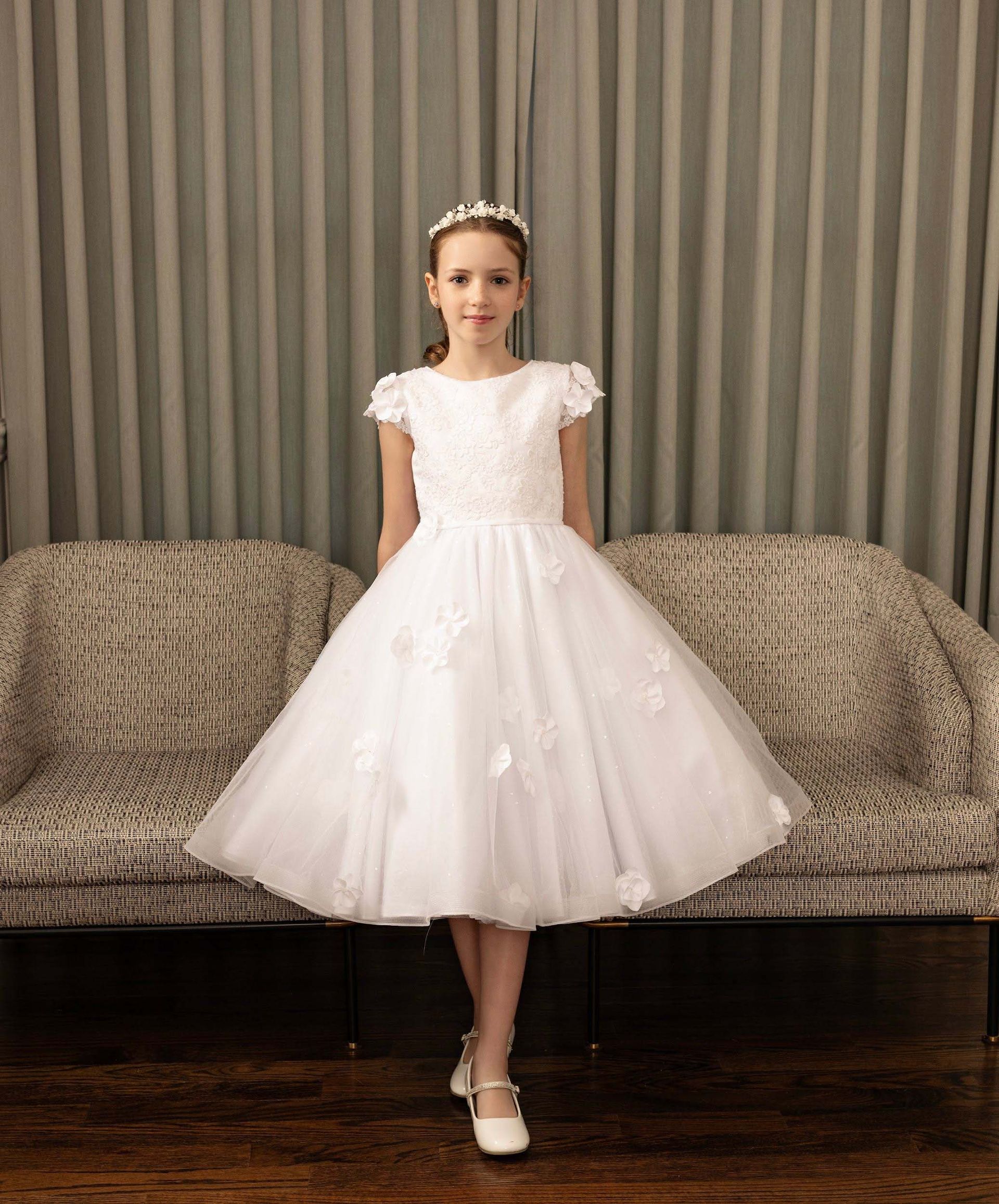 Child in a white dress and tiara standing in front of a couch and curtains