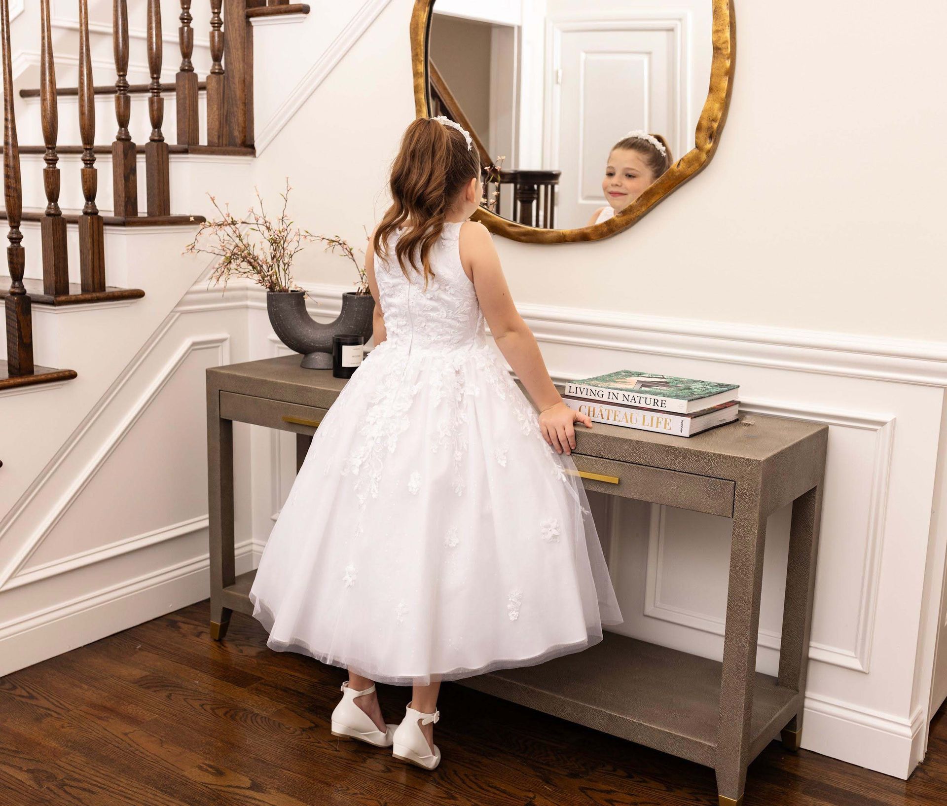 Girl in a white dress looking into a round mirror beside a console table and staircase.