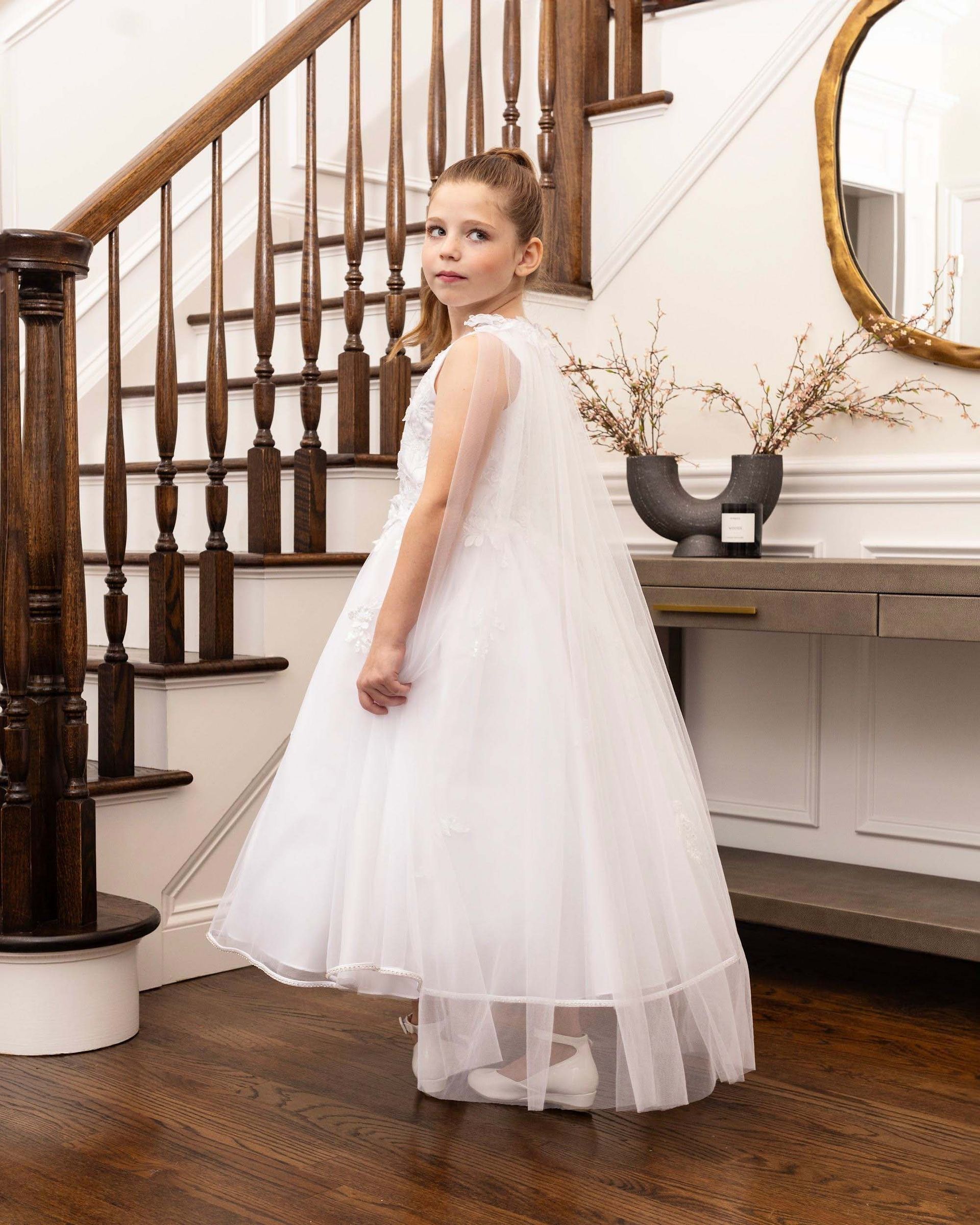 Girl in a white tulle dress standing by a staircase in a bright hallway, looking back over her shoulder