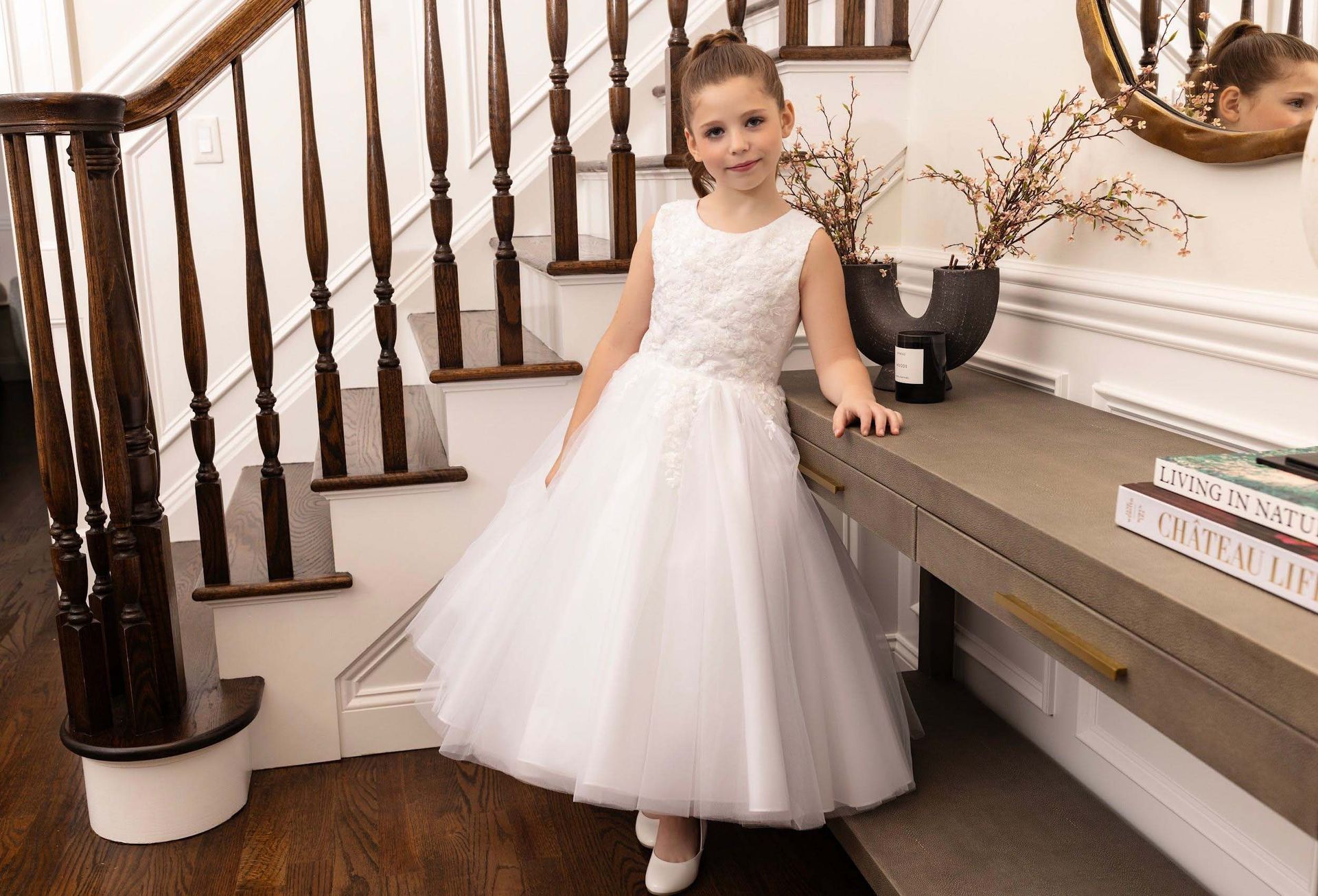 Young girl in a white dress standing by a staircase and console table in a bright room