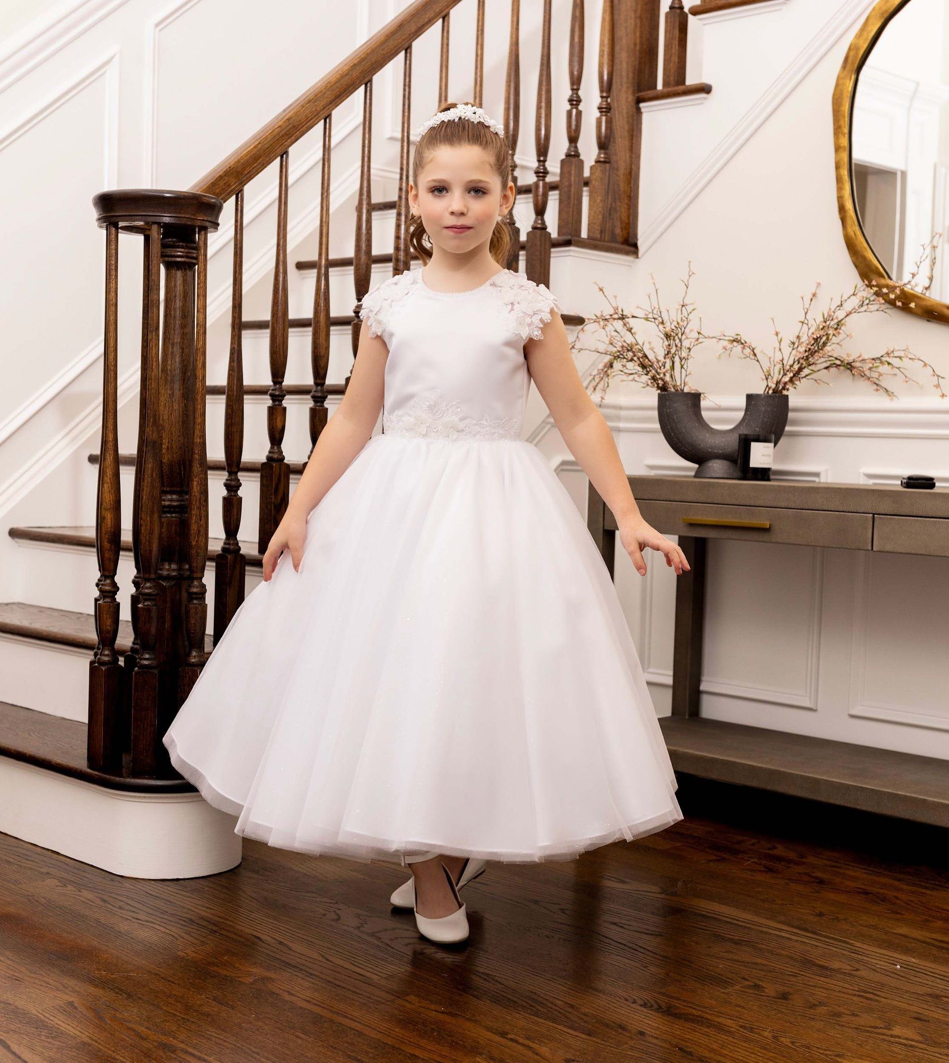 Girl in a white dress standing in a staircase foyer, posing on a wooden floor.