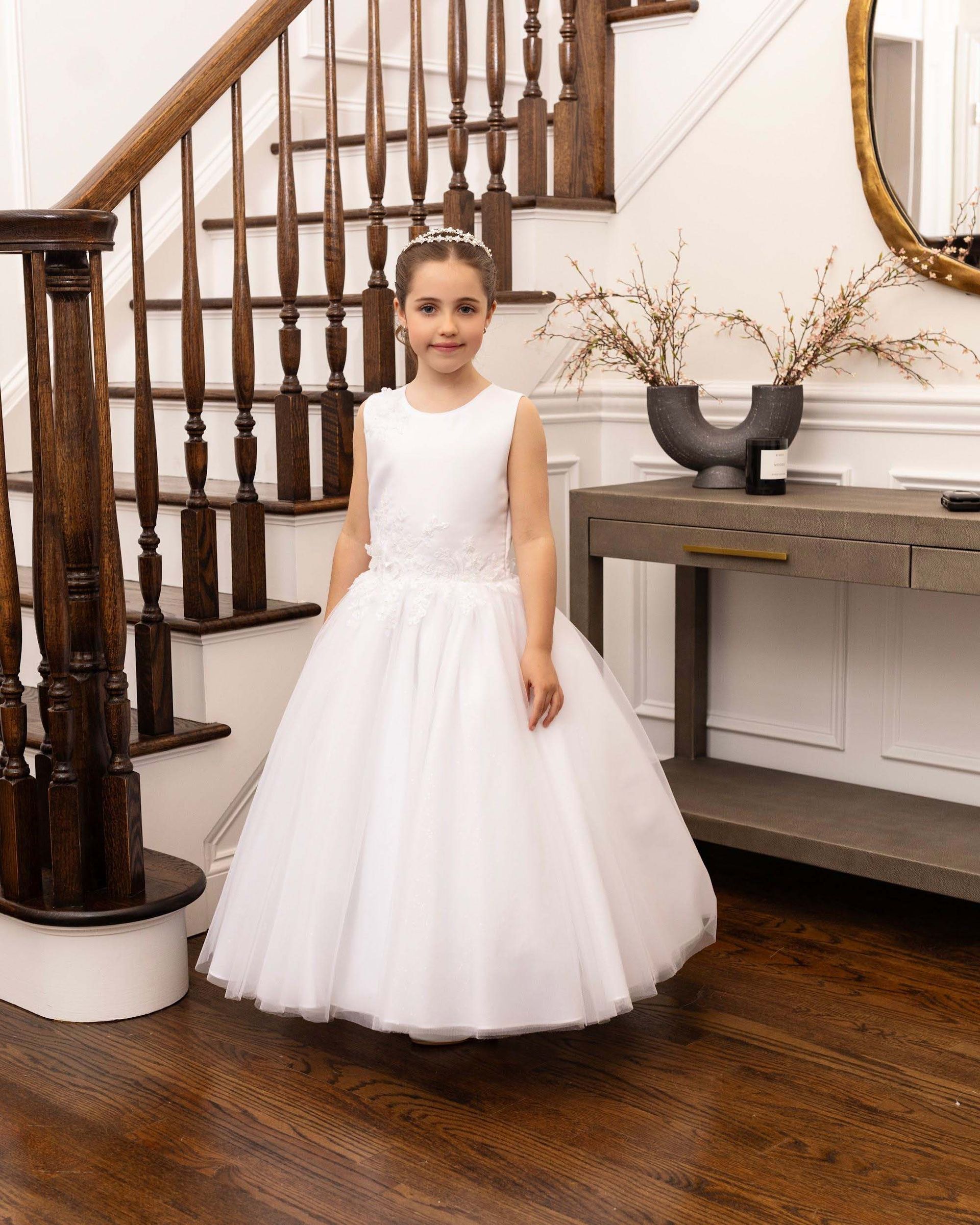 Child in a white formal dress standing in a bright home interior by a wooden staircase