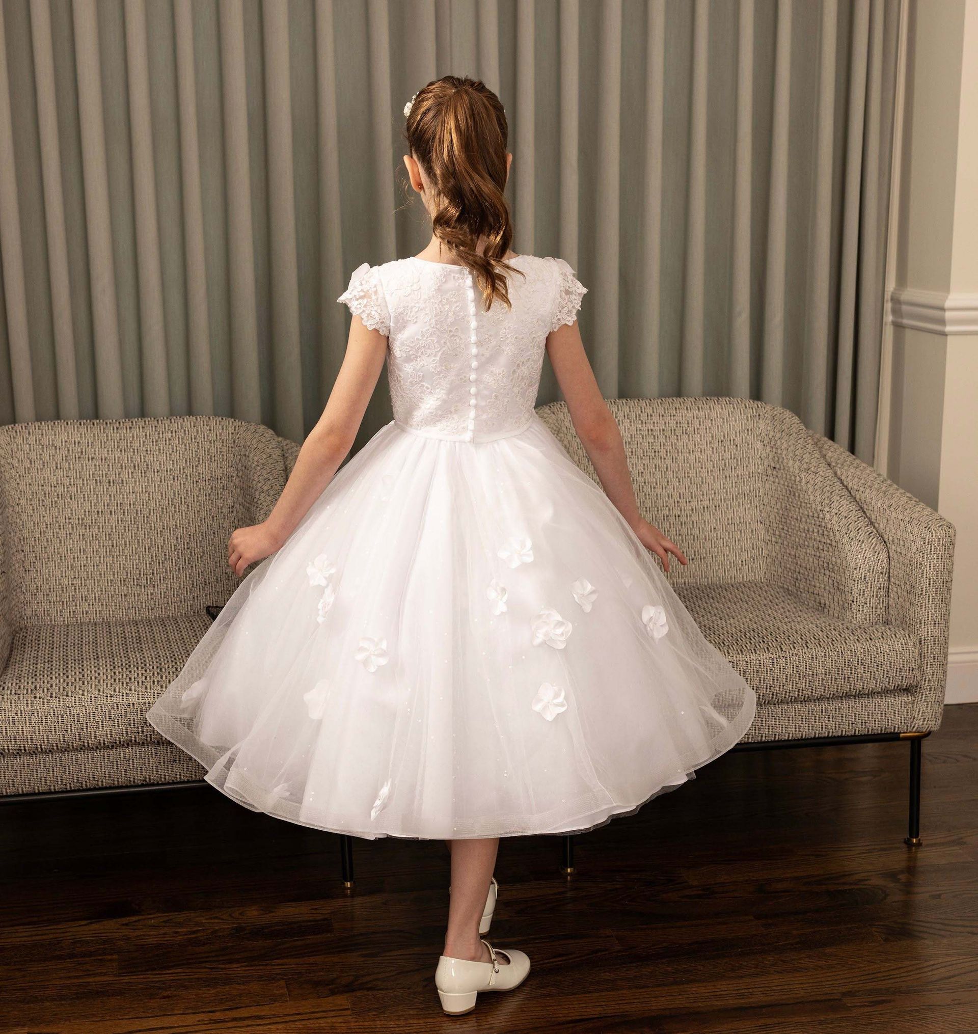Girl in white dress standing in a living room with her back to the camera, holding the hem of her skirt