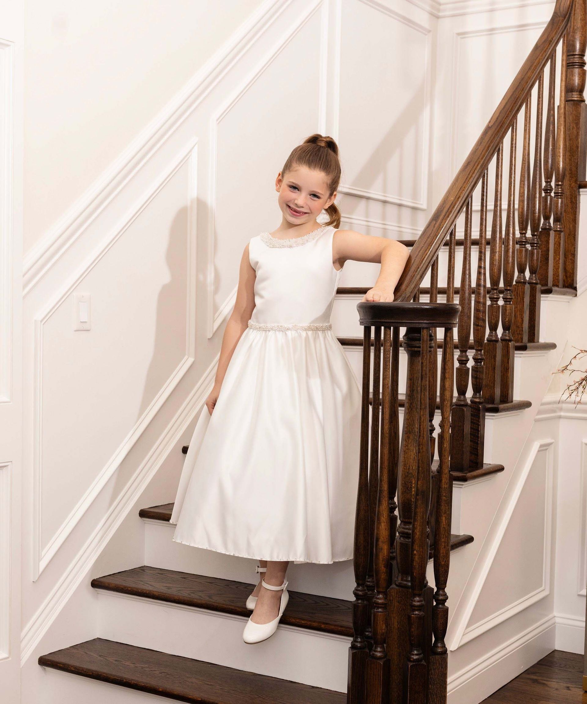 Child in a white dress standing on a staircase with dark wood railings and white walls.