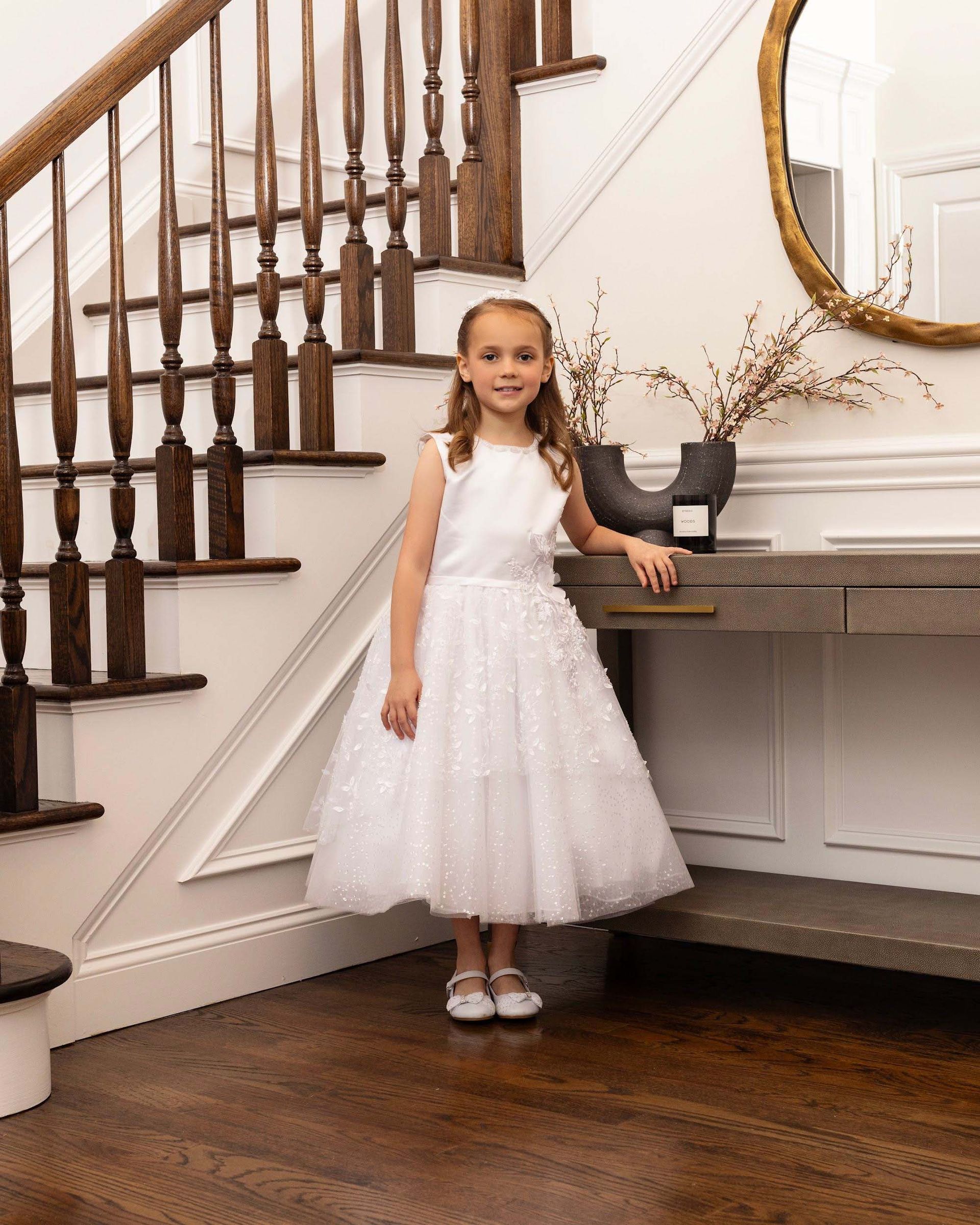 Girl in a white dress standing by a staircase and console table in a bright interior.