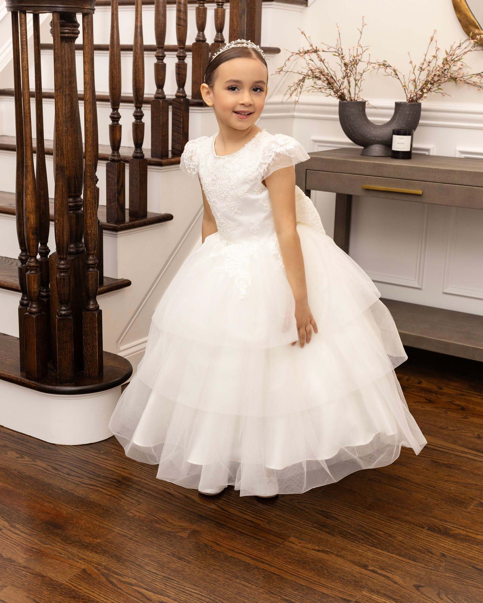 Young child in a white formal dress standing indoors on a wooden floor near a staircase.