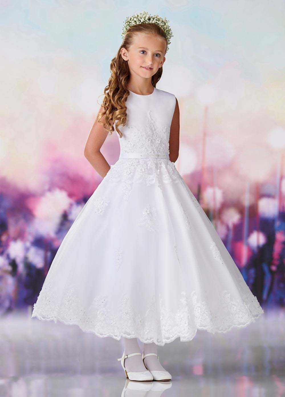 Young girl in a white formal dress and floral headband, standing against a soft pastel background.