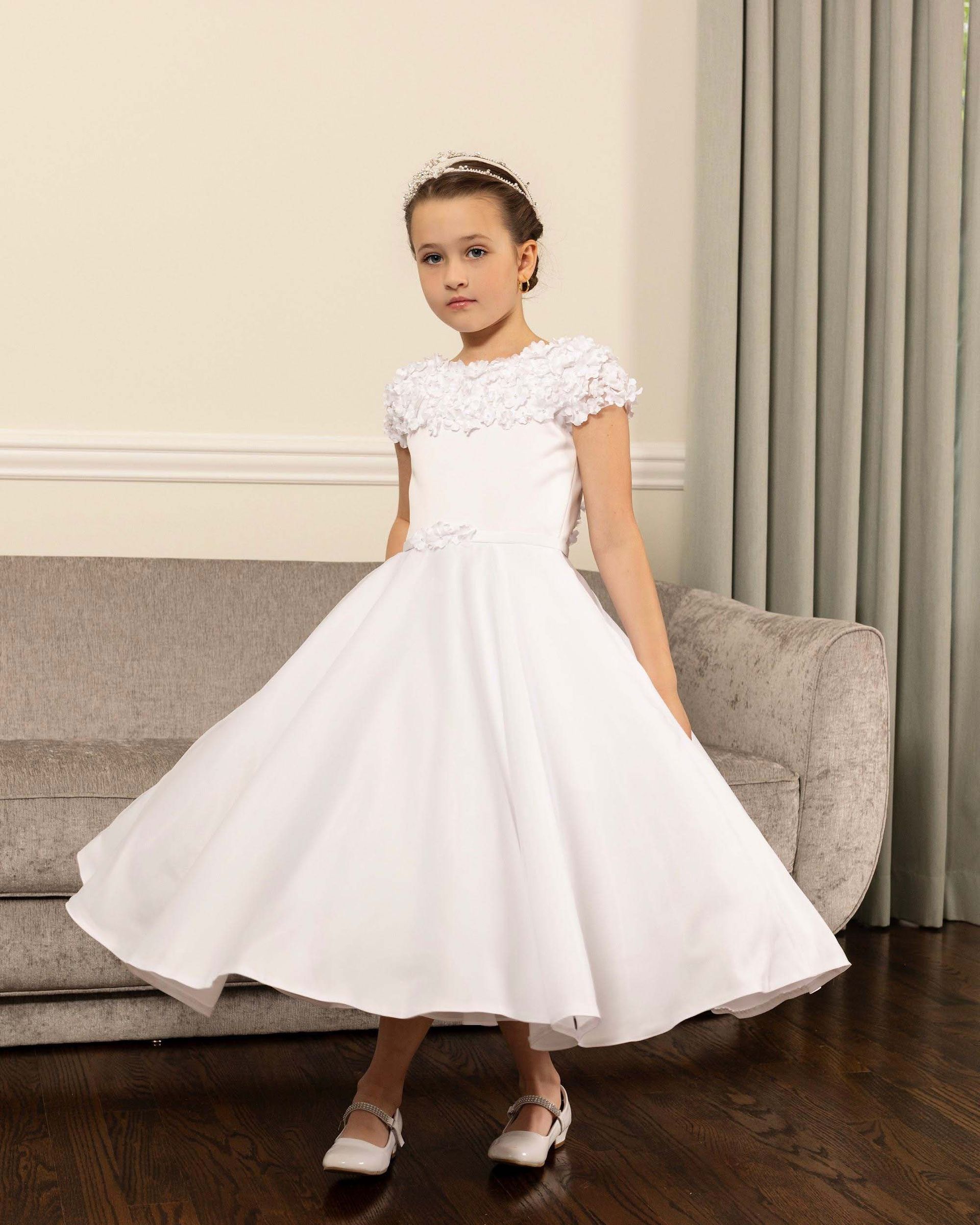 Child in a white dress standing indoors against a beige sofa and curtains