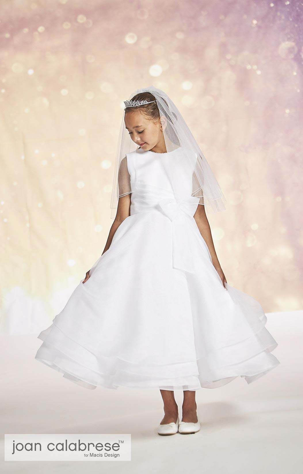 Young child in a white dress and veil posing in a studio with a soft pink, sparkly backdrop