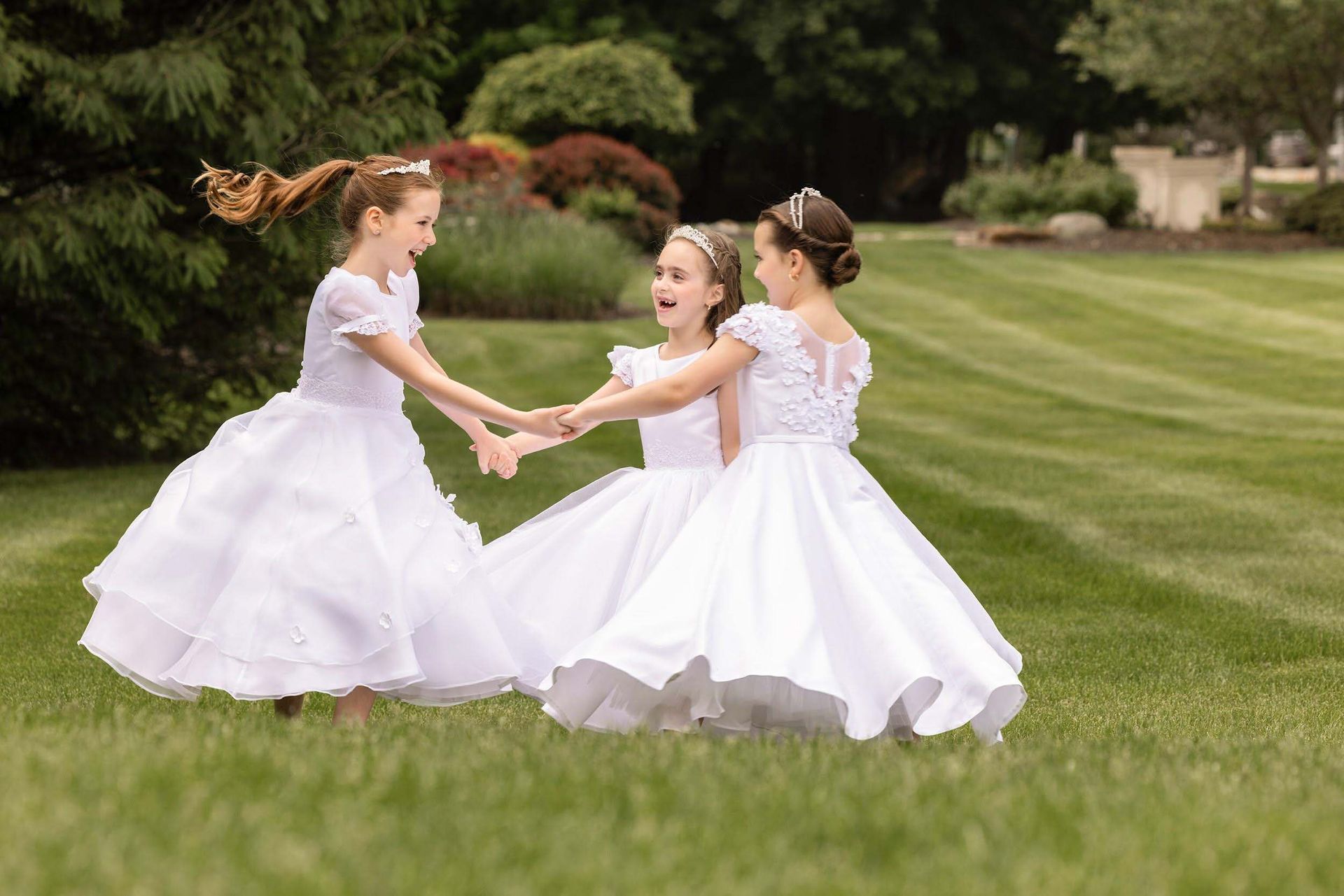 Three girls in white dresses holding hands and spinning on a grassy lawn