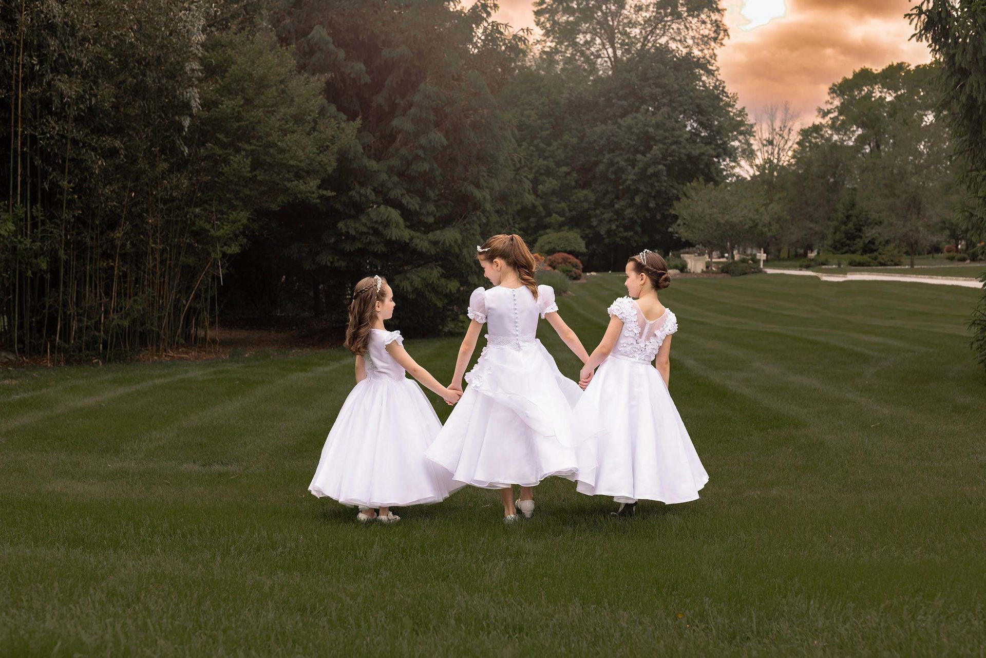 Three girls in white dresses standing on a grassy lawn at sunset