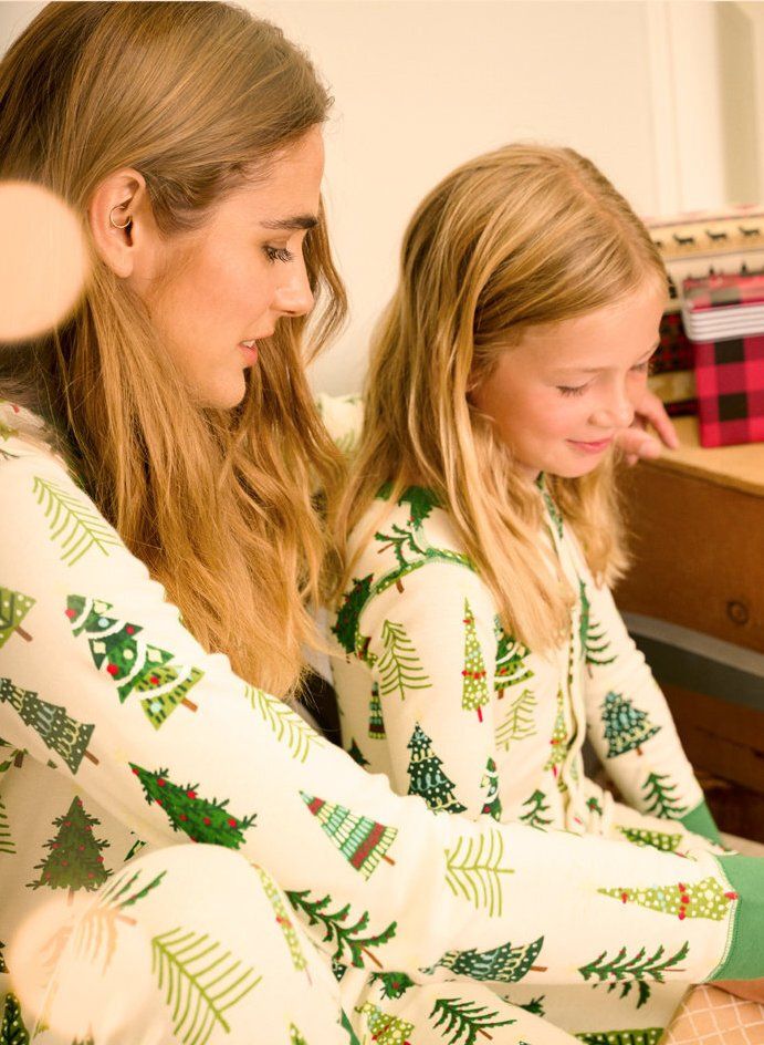 Two children in matching white pajamas with green trees sit together indoors, looking down and smiling.