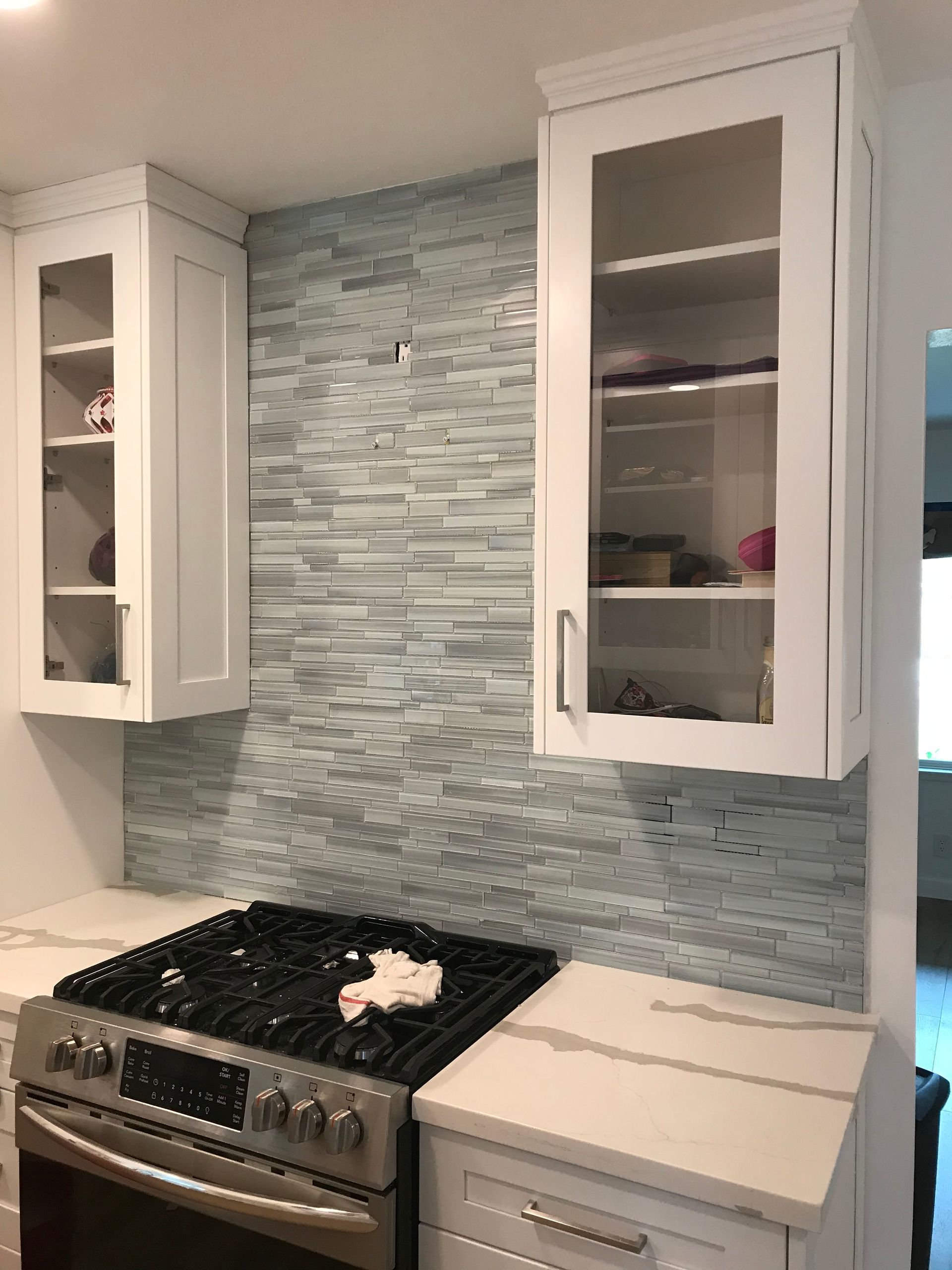 Kitchen stovetop with white cabinets and gray tile backsplash, one cabinet door open.
