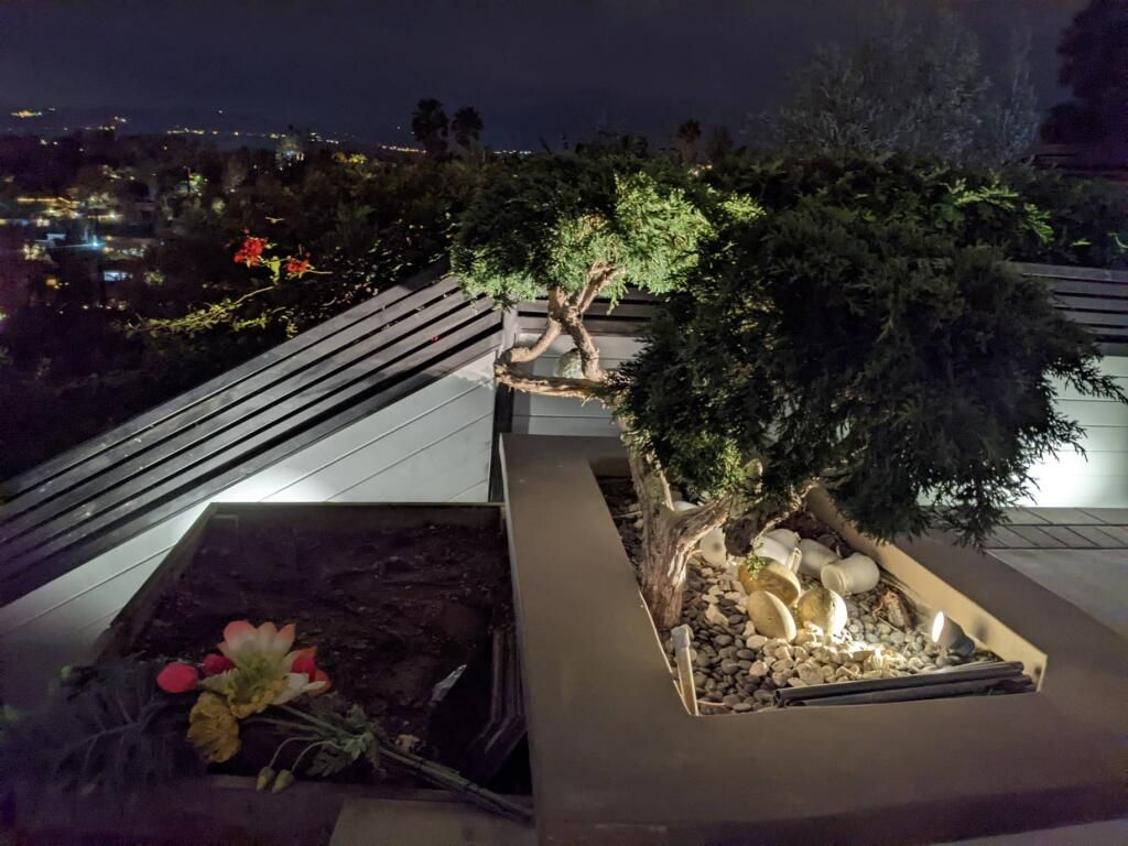 Night rooftop garden with lit planters, a small statue, and city lights in the background