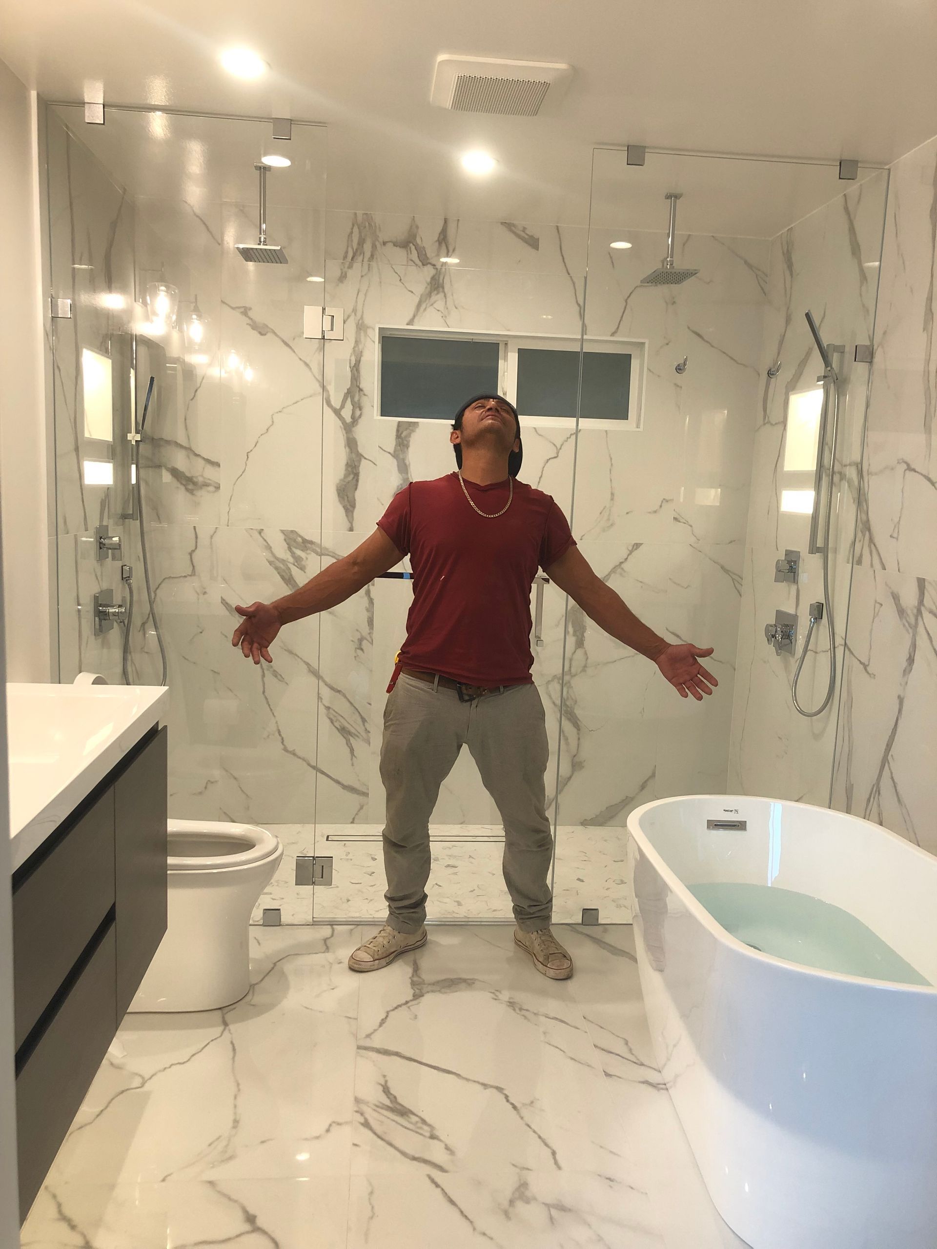 Person standing with arms outstretched in a marble-tiled bathroom beside a bathtub and toilet
