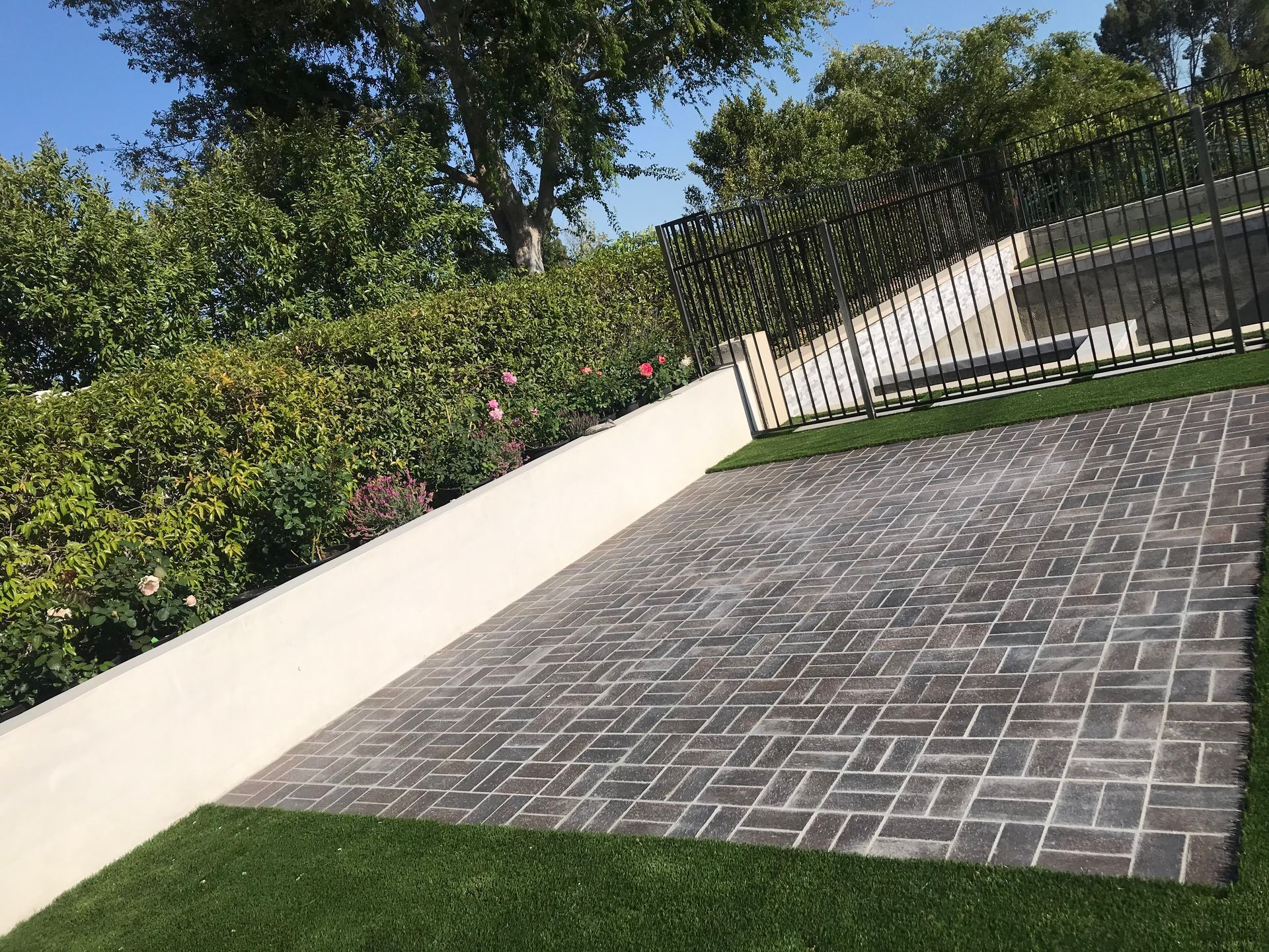Brick-paved driveway beside a white retaining wall and black fence, with grass and trees in the background