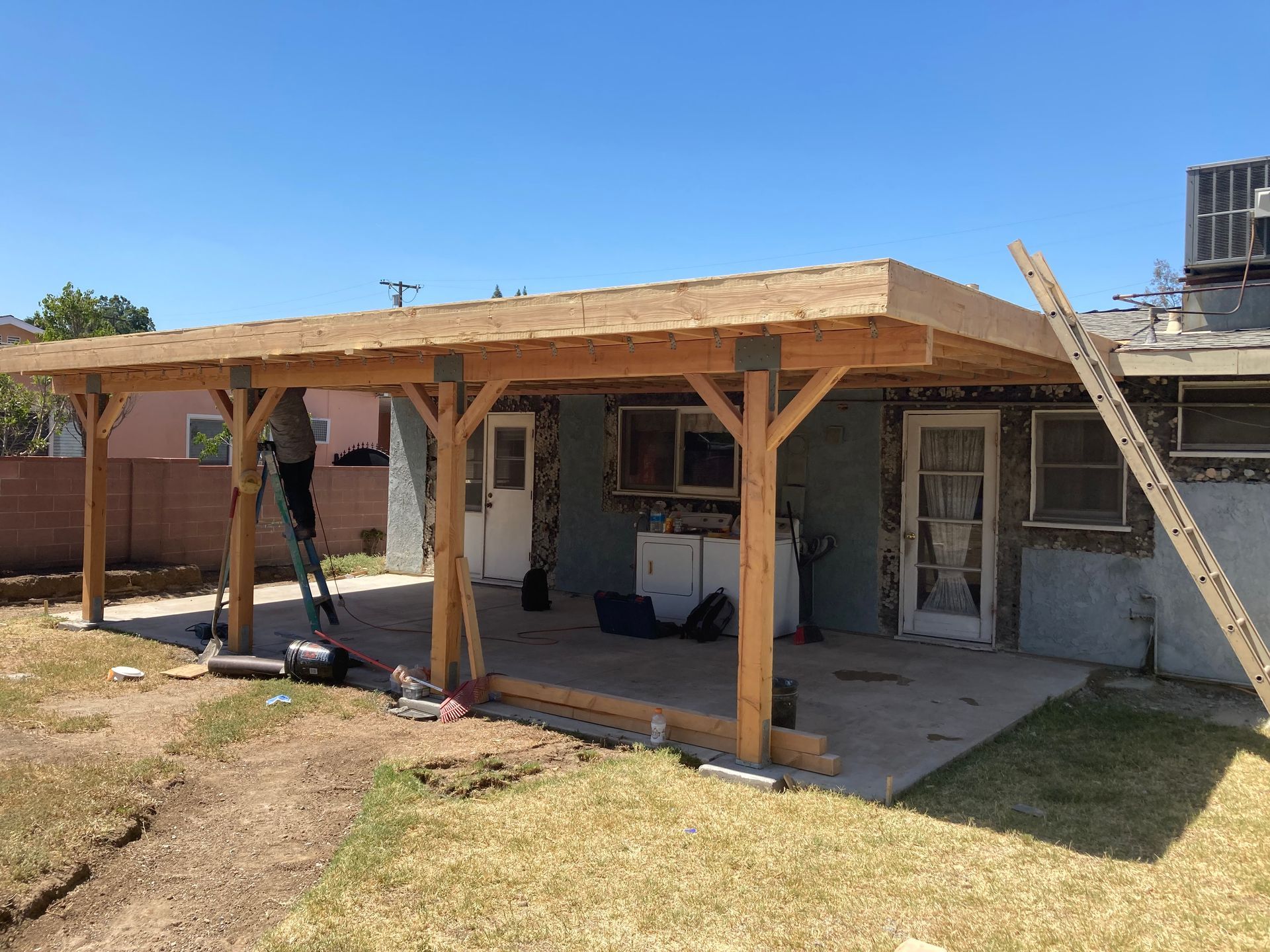 Back patio with a new wooden pergola roof attached to a house, over a concrete slab and lawn.