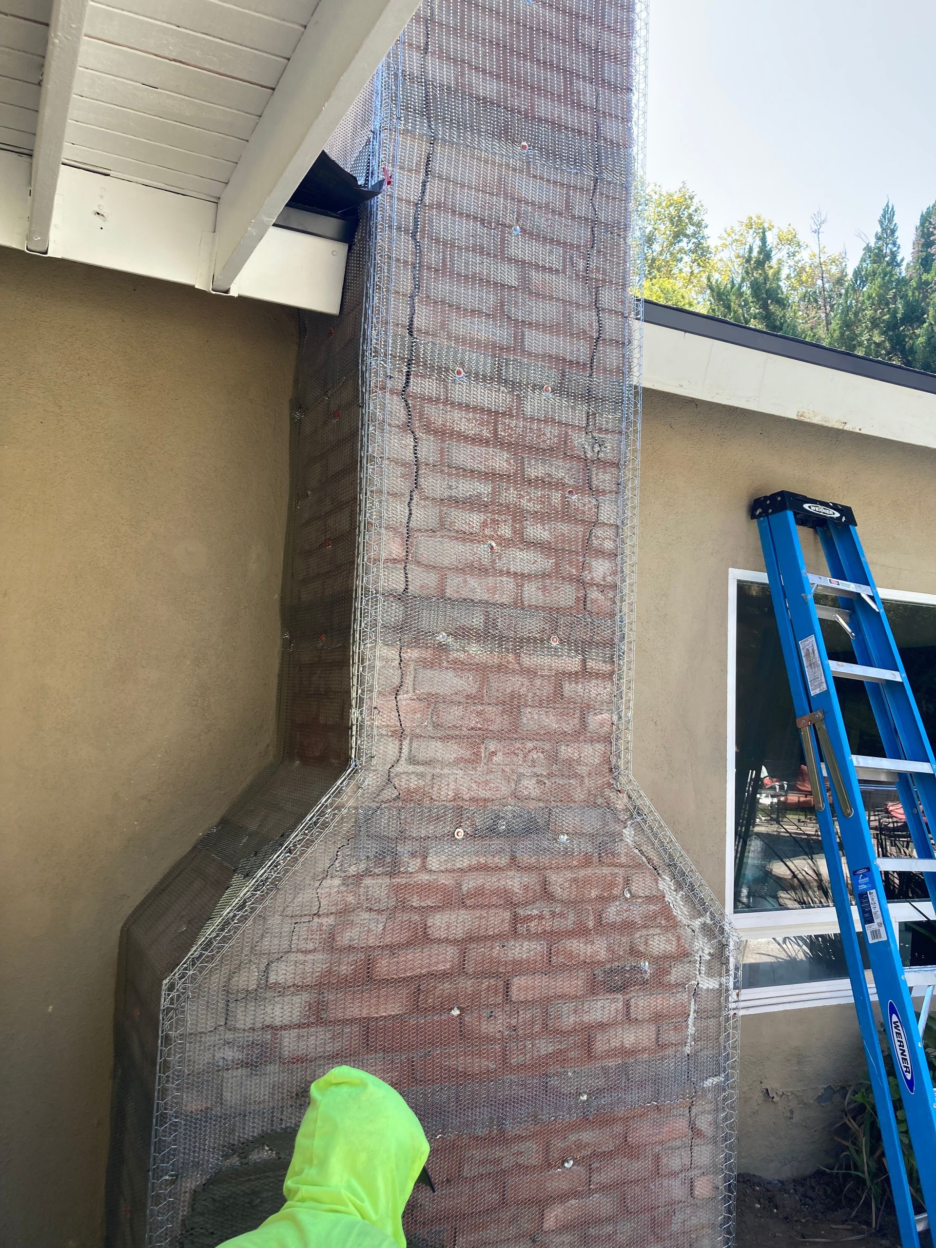 Brick chimney under a covered patio, with a blue ladder beside it and a person in a green hooded jacket below.