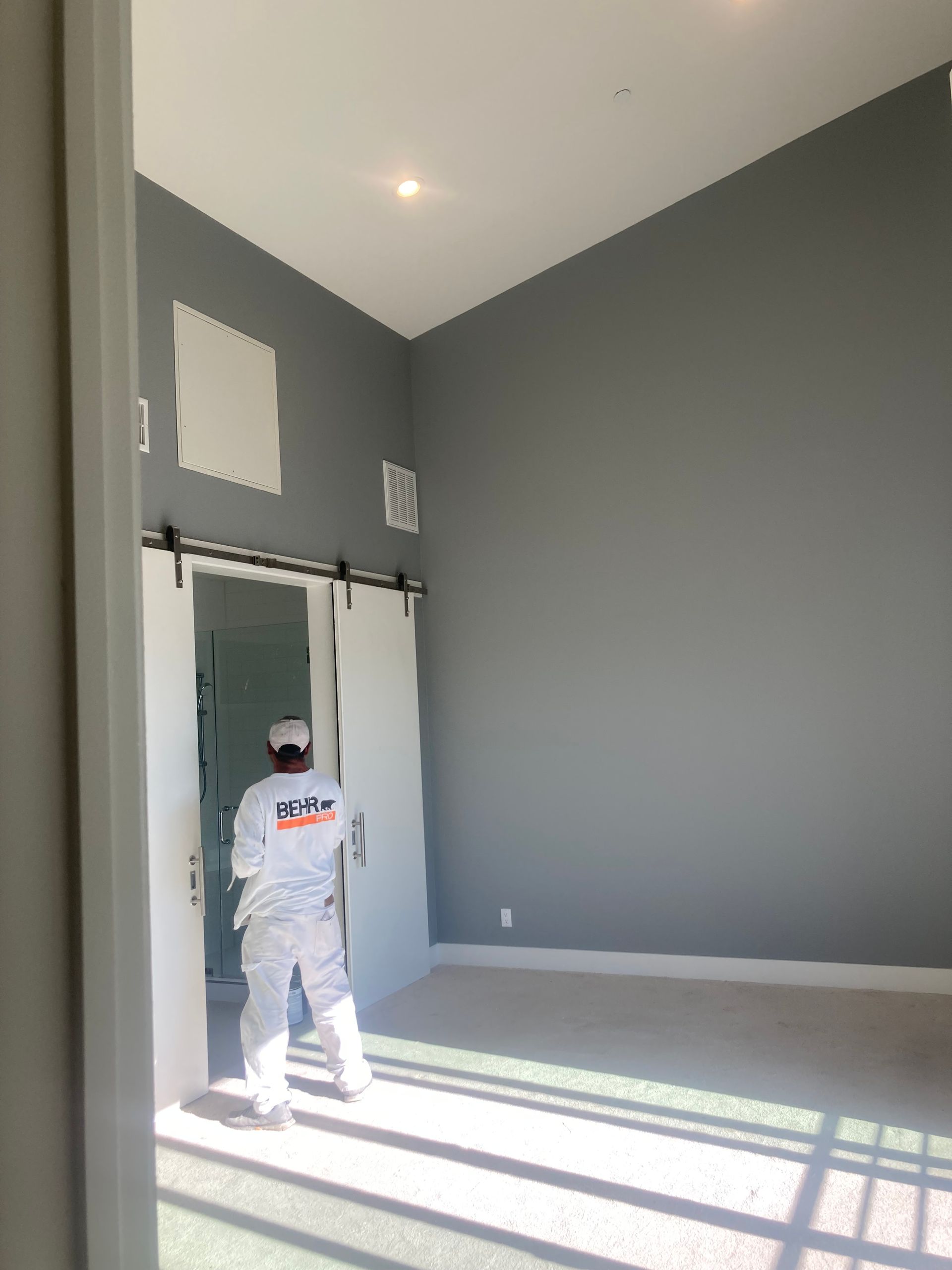 Worker painting a tall gray room with white trim and sunlight streaming through large windows.