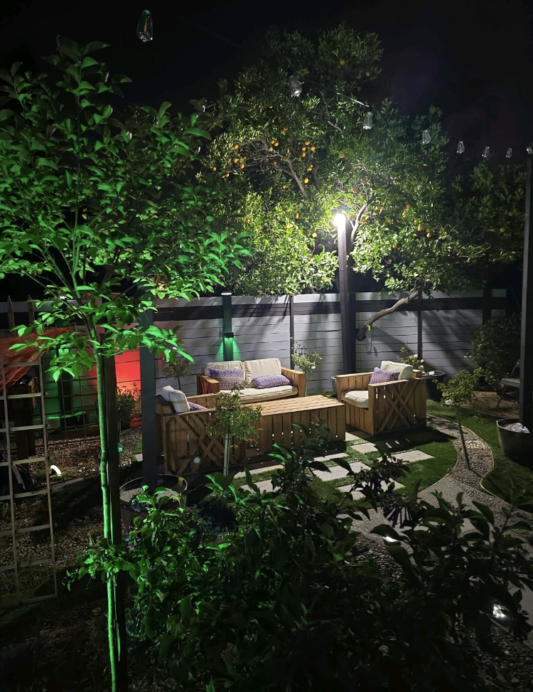 Nighttime garden patio with lit seating area, trees, and a lamppost behind a fence.